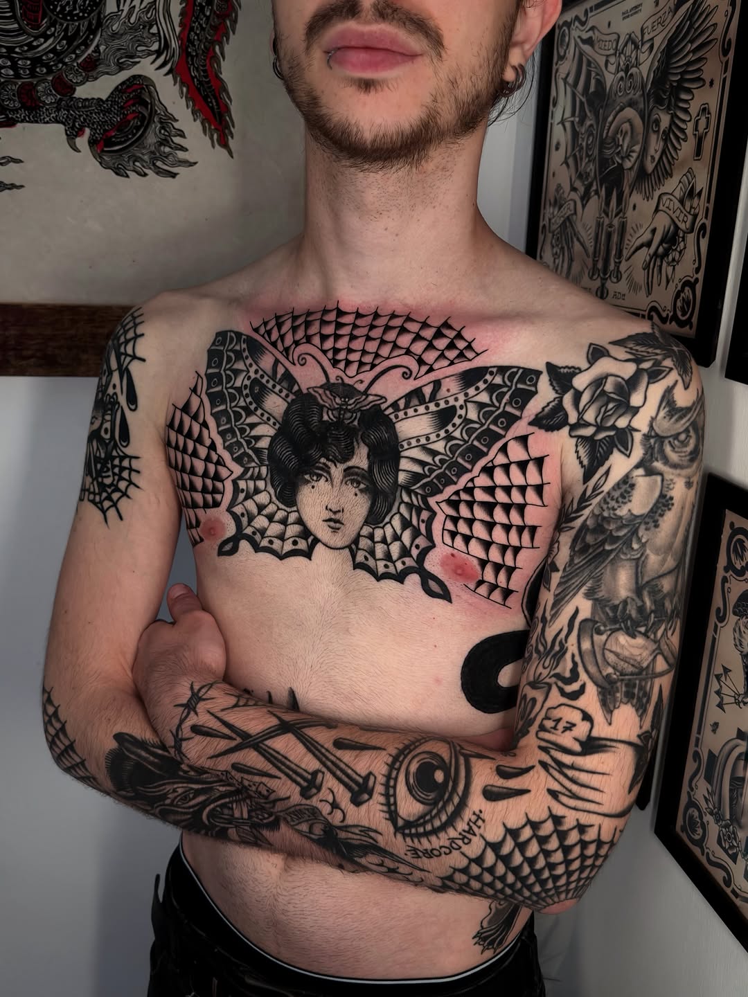 Chest and arm tattoos featuring a winged female face with ornamental scales, florals, and decorative linework in black ink.