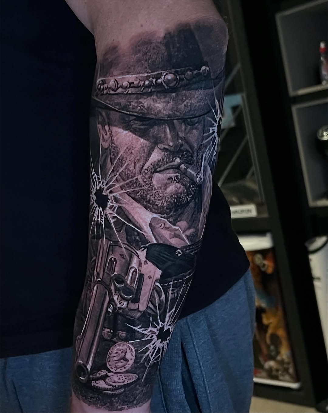 Realistic black and grey sleeve tattoo featuring a bearded man with ornate headpiece, revolver, sunburst, coins, and decorative elements on upper arm.