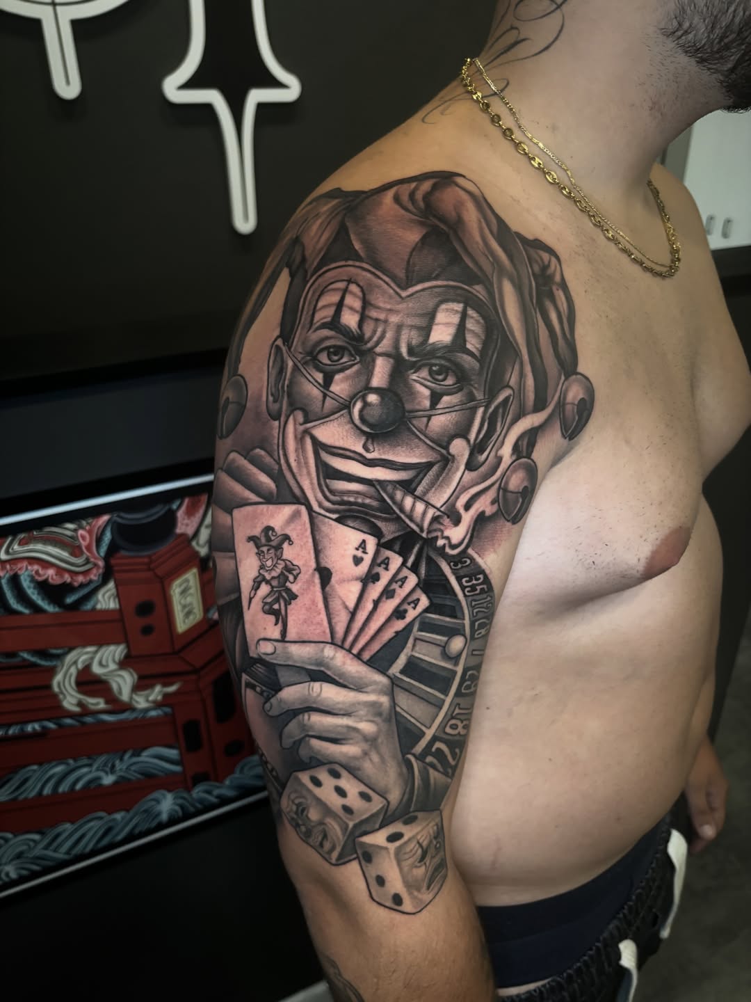 Joker face tattoo with playing cards and dice on shoulder, rendered in black and grey with smooth shading.