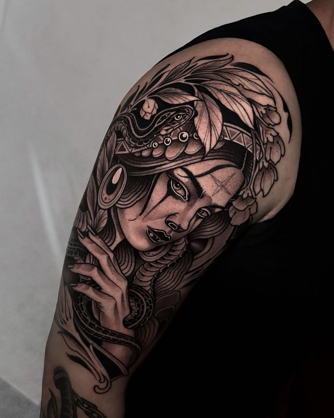 Ornate black and grey portrait of a woman's face with elaborate feathered headdress and decorative elements on upper arm.