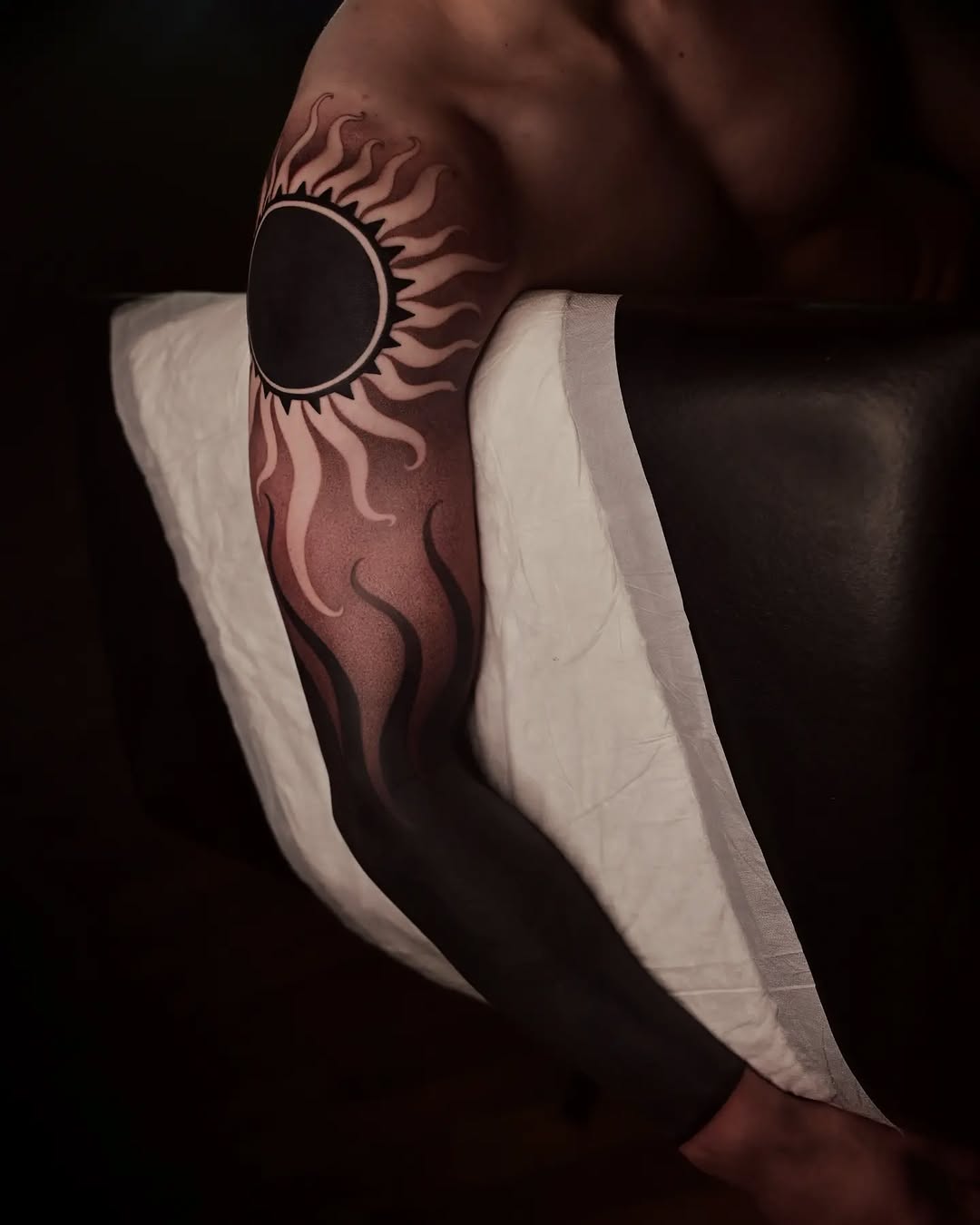 Sun with radiating flame-like rays in black and grey shading on upper arm, wrapped in white bandage.
