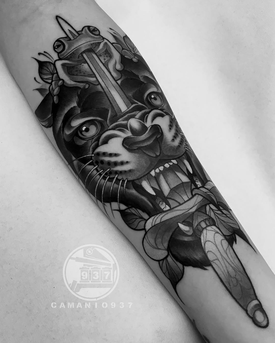 Black and grey arm tattoo featuring a frog with ornate mask-like facial markings, surrounded by decorative insects and foliage in smooth shading.