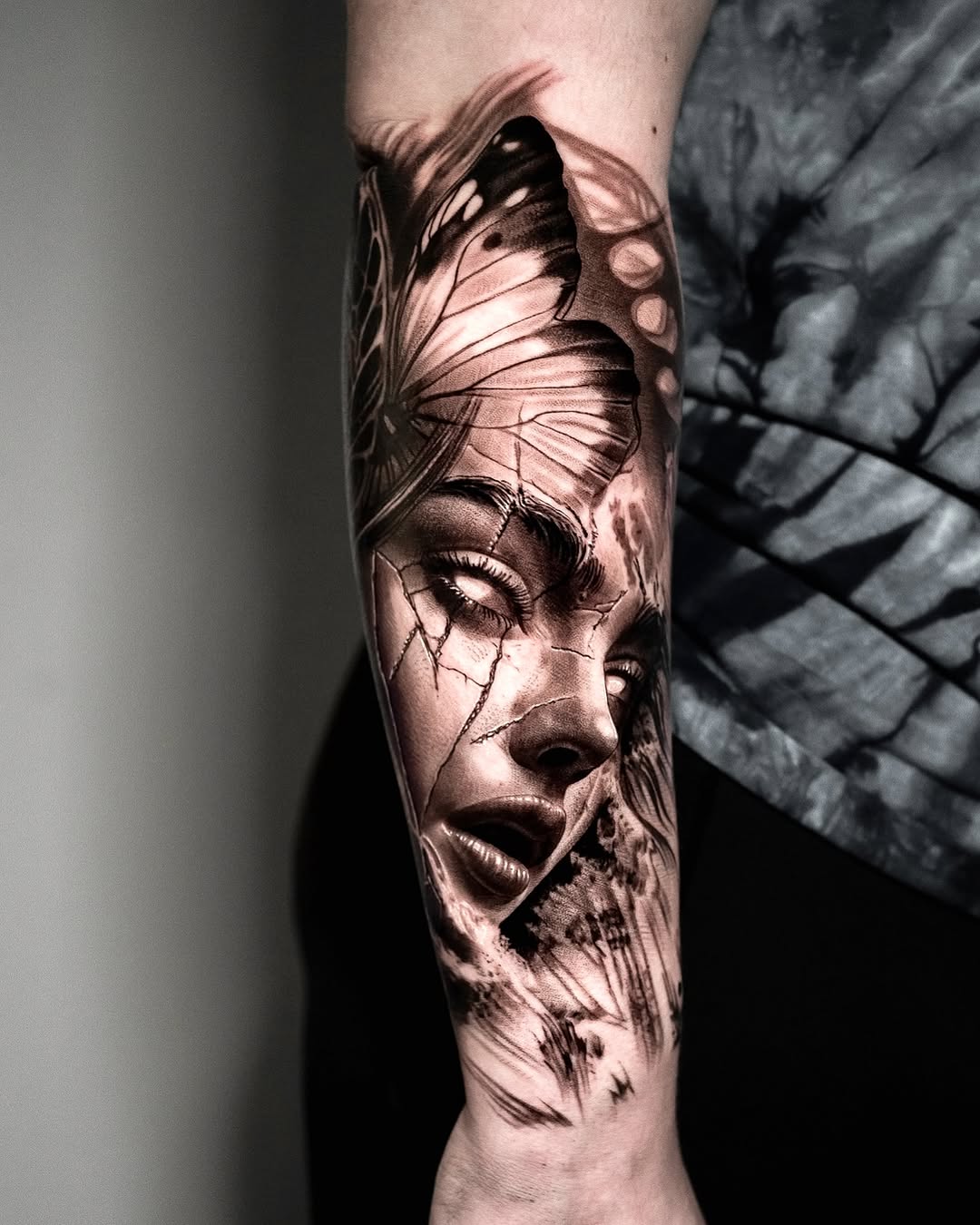 Woman's forearm featuring a black and grey portrait of a face with butterfly wings, rendered with soft shading and realistic details.