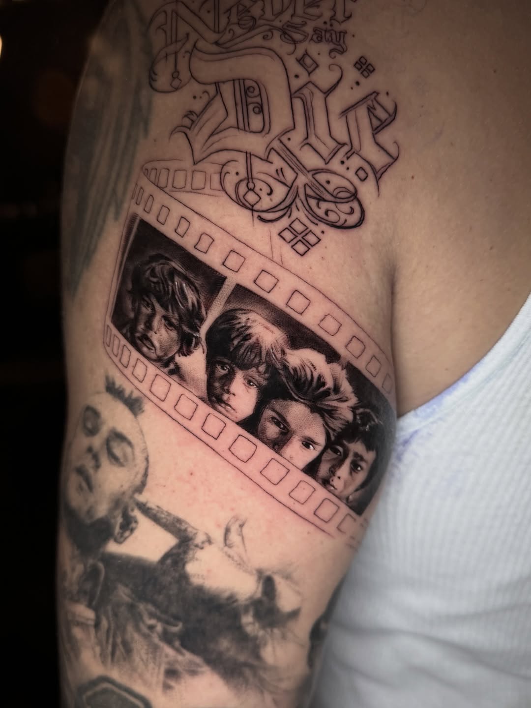 Black and grey arm tattoo featuring decorative line work at top and a film strip with four portrait faces below, positioned on upper arm.