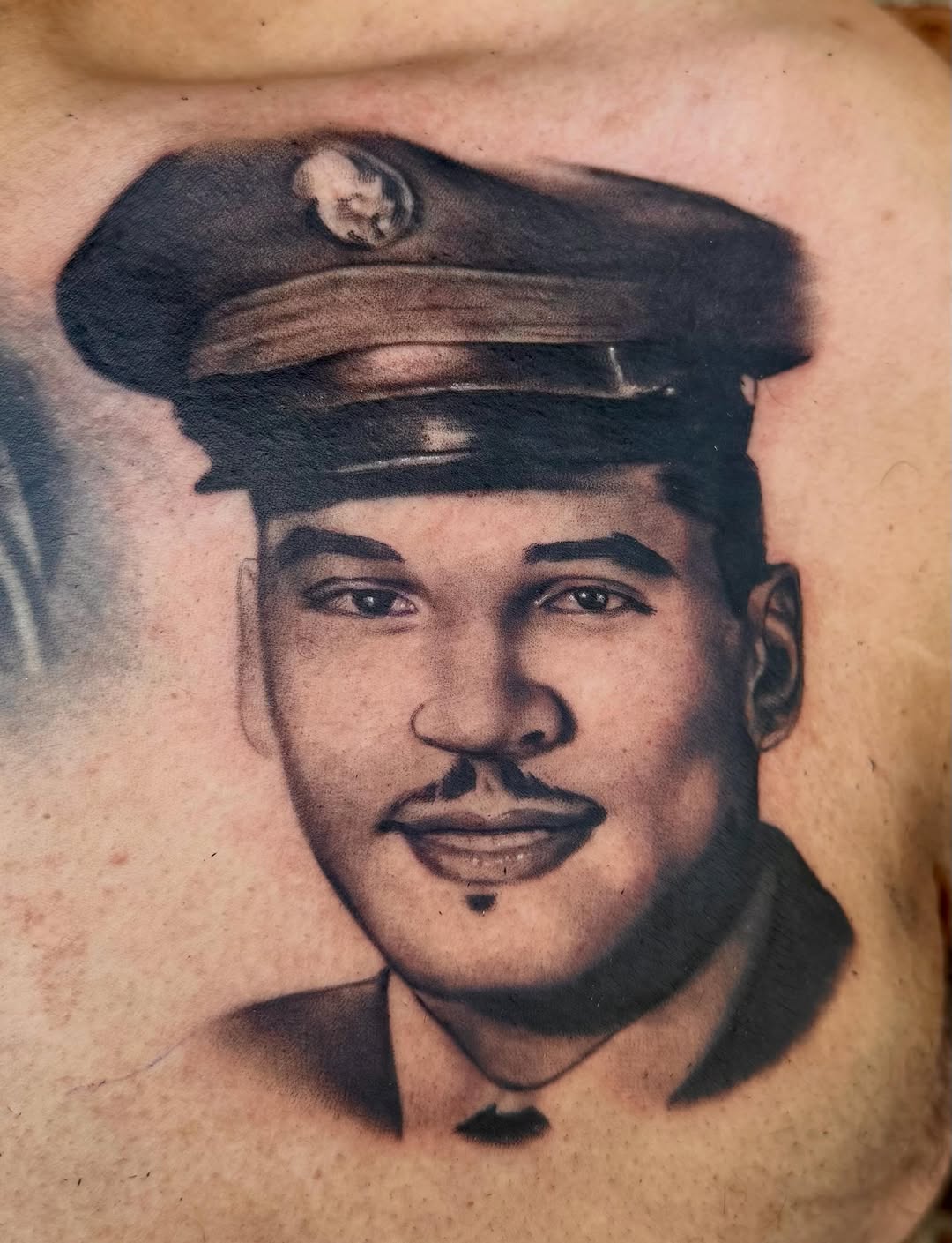 Portrait tattoo of a man wearing a military cap with smooth black and grey shading, located on upper arm or chest.