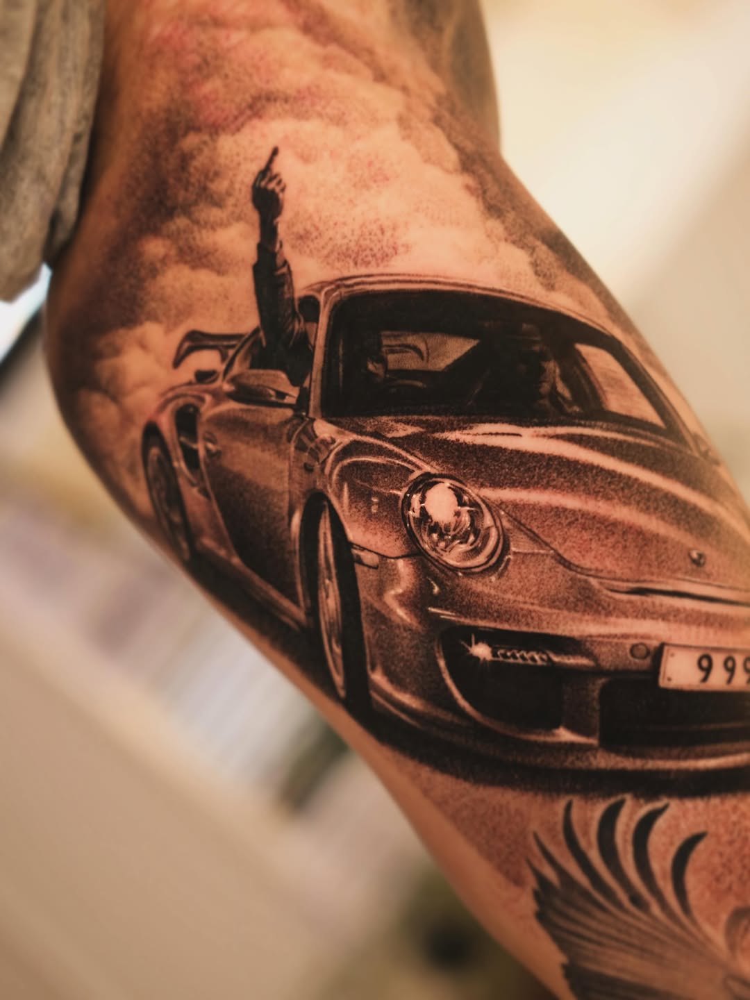 Black and grey realistic car tattoo on upper arm with a figure on the roof and license plate visible.