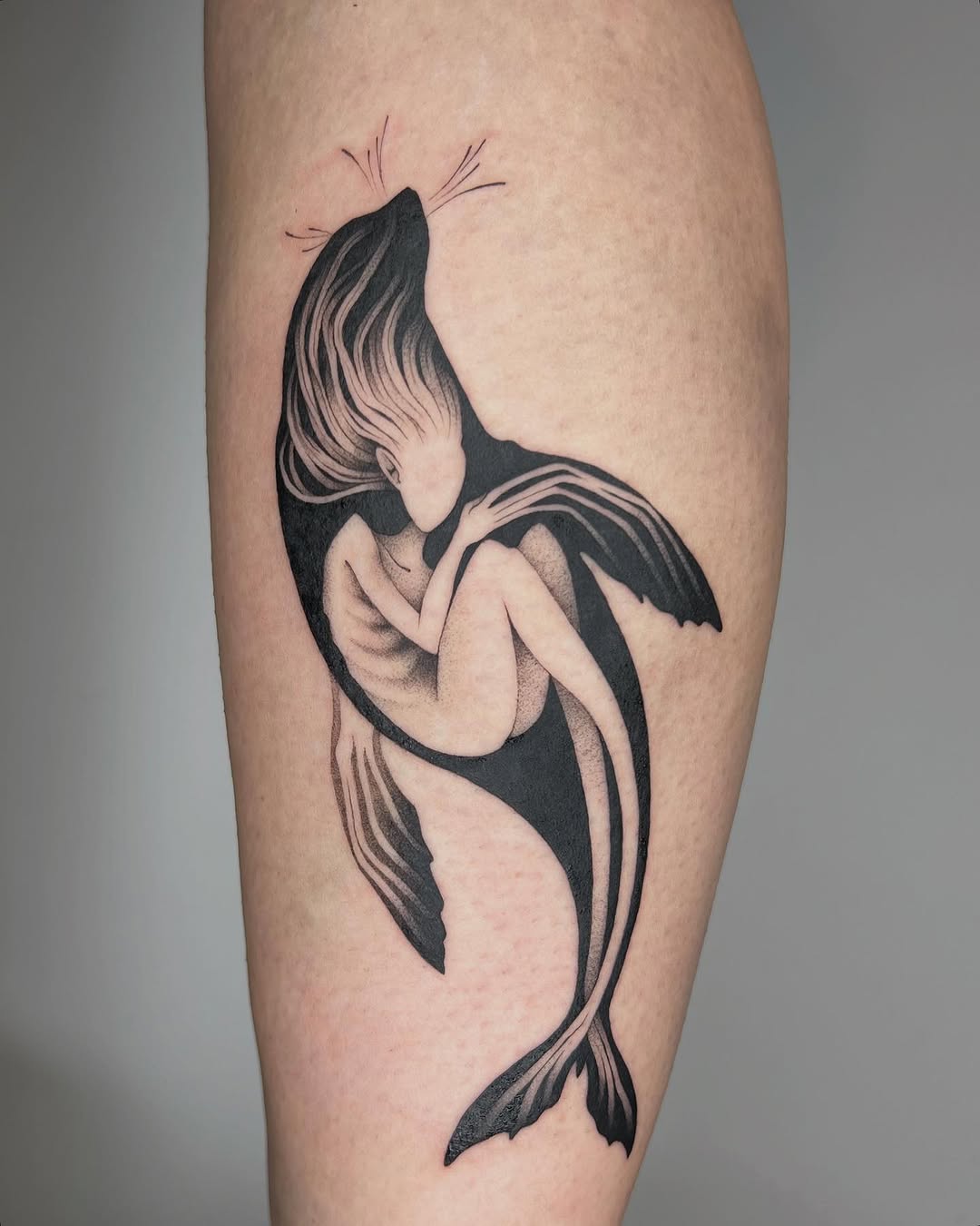 Mermaid tattoo with flowing hair and striped tail on calf in black and grey with soft shading.