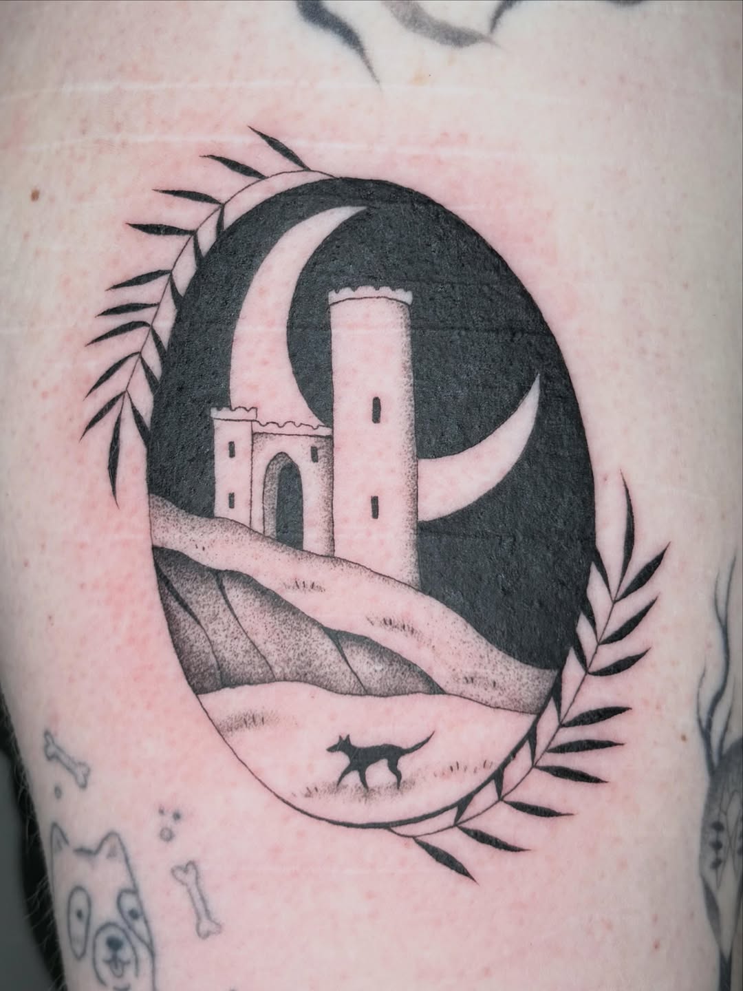 Black and grey tattoo on upper arm featuring a crescent moon with a tower and castle, surrounded by decorative botanical elements and a small fox silhouette.