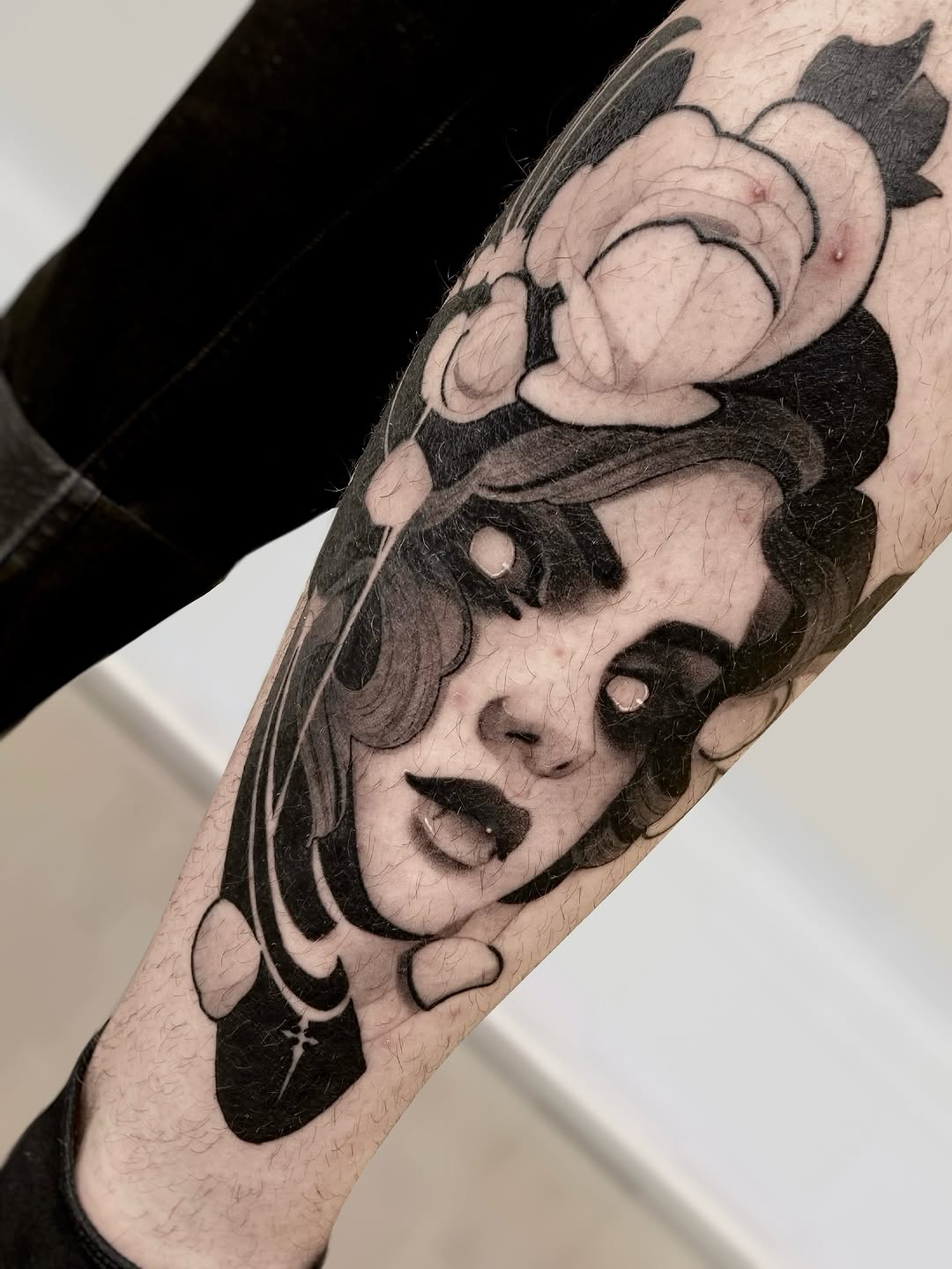 Black and grey portrait tattoo on forearm featuring a woman's face with dramatic expression and decorative elements above, rendered with soft shading.