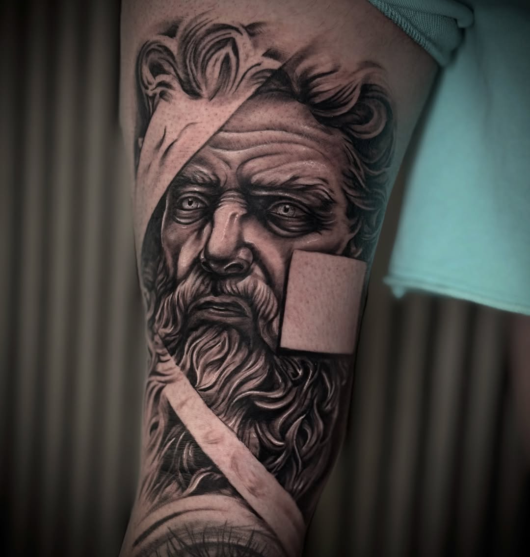 Black and grey portrait tattoo of a bearded man with curly hair on upper arm, featuring detailed facial shading and realistic features.
