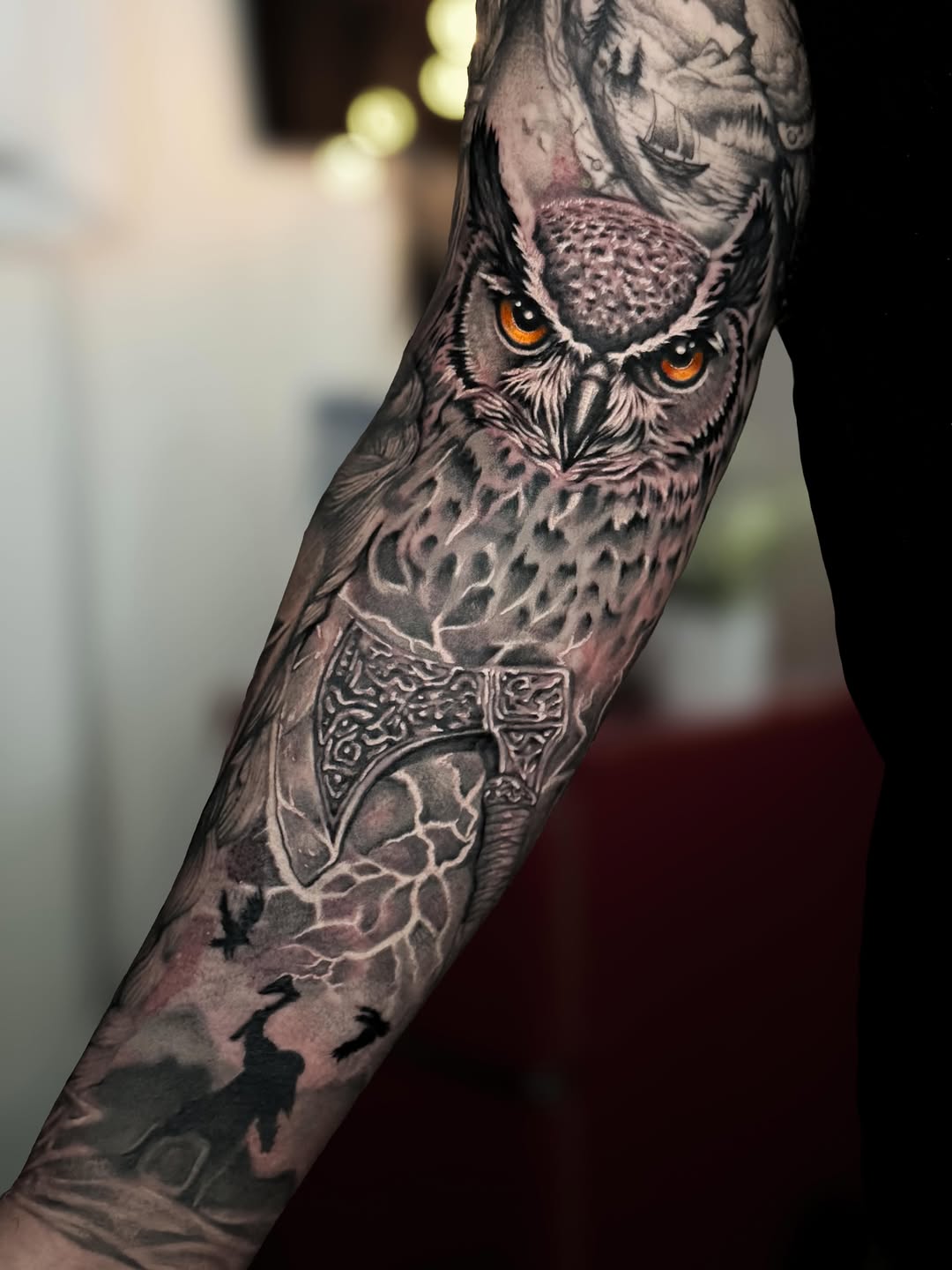 Owl with striking amber eyes and detailed feathers on forearm, featuring ornate Celtic axe design and silhouetted figures below in black and grey.