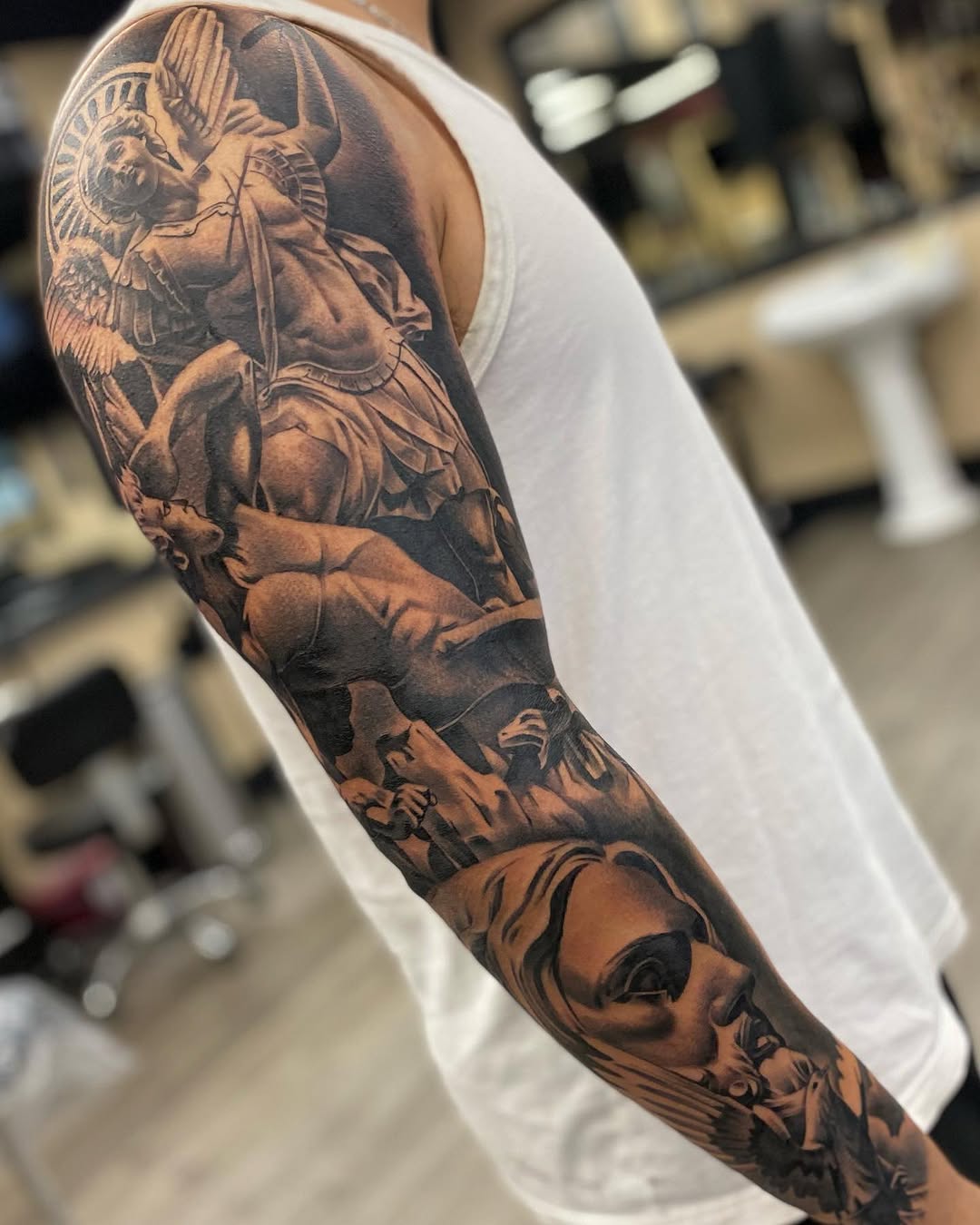 Saint Michael Archangel full sleeve tattoo with detailed armor and wings in black and grey shading on muscular arm.