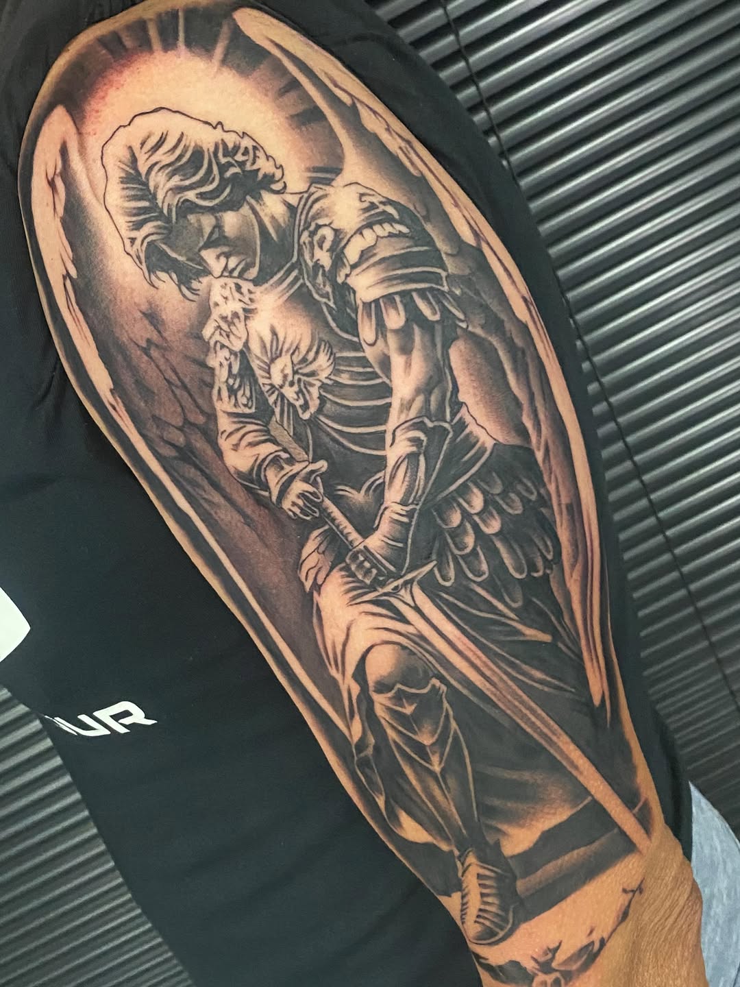 Saint Michael the Archangel with wings and armor defeats a demon on upper arm, rendered in black and grey with smooth shading.