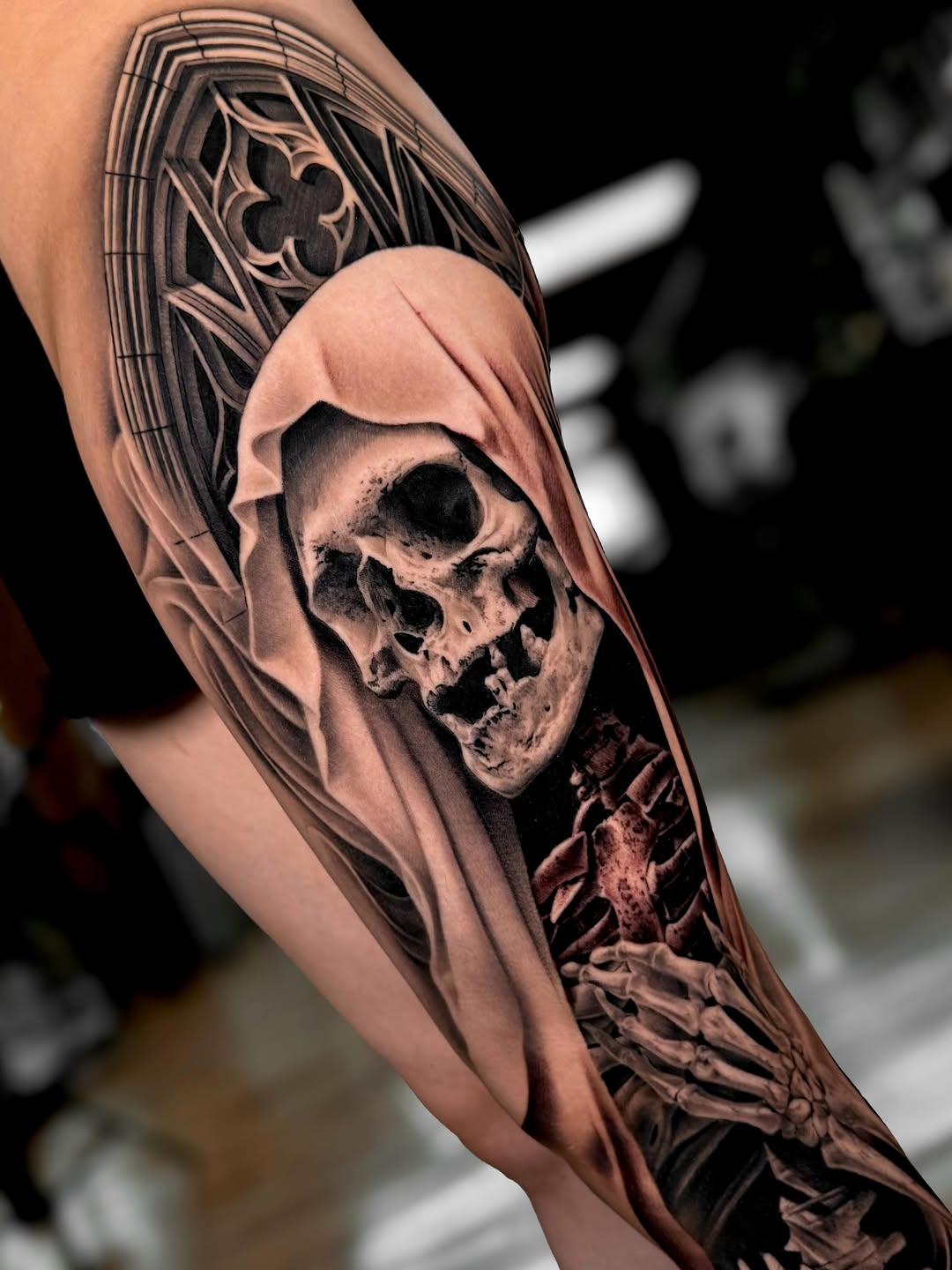 Reaper figure with skull face wearing hooded robes on forearm, with ornate geometric upper arm panel, black and grey shading.