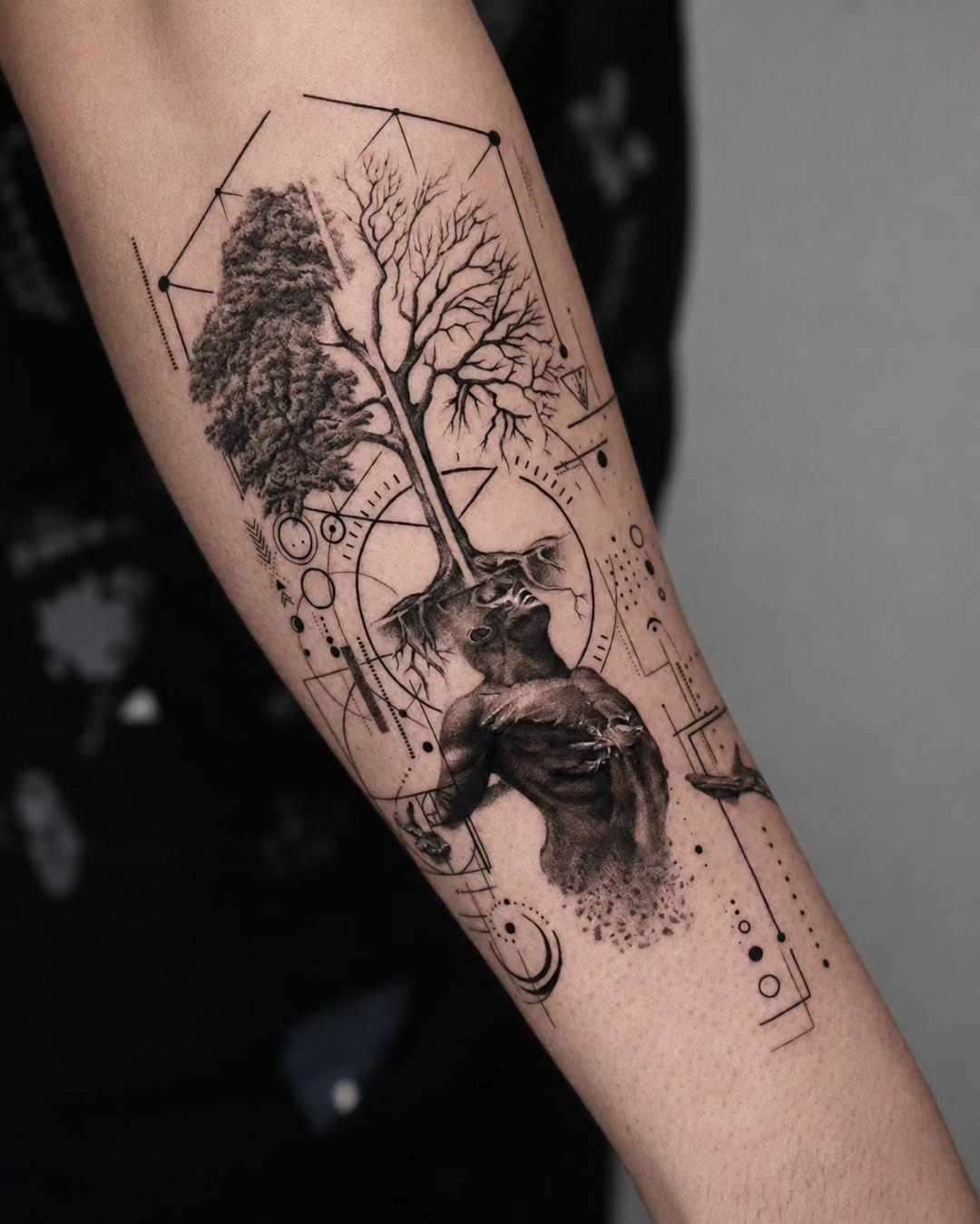 Black and grey tattoo on forearm featuring a tree with detailed roots, geometric line art, and an animal head portrait below.