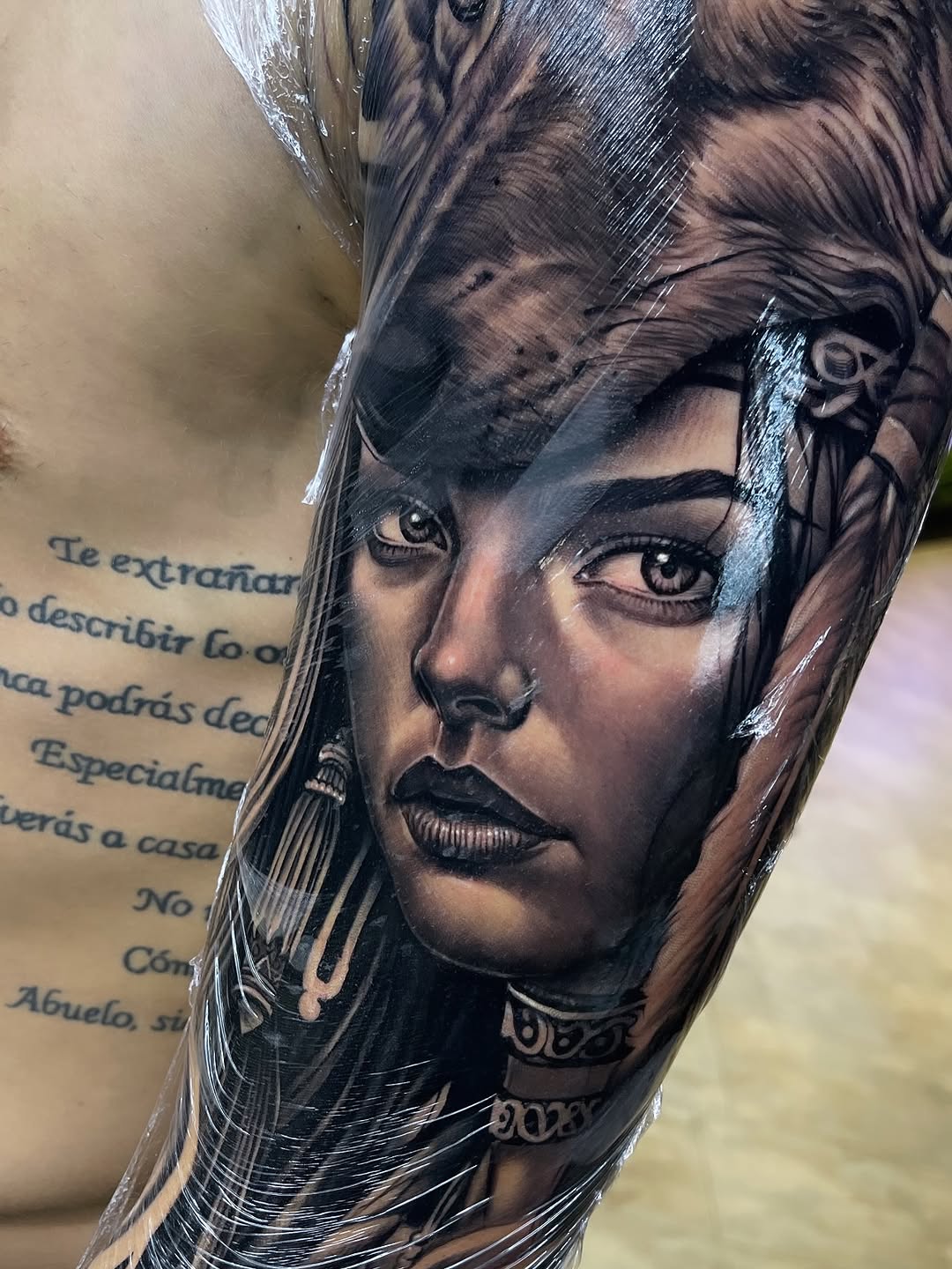 Black and grey portrait tattoo of a woman's face with dramatic eye detail and flowing hair on an arm, with Spanish text visible nearby.