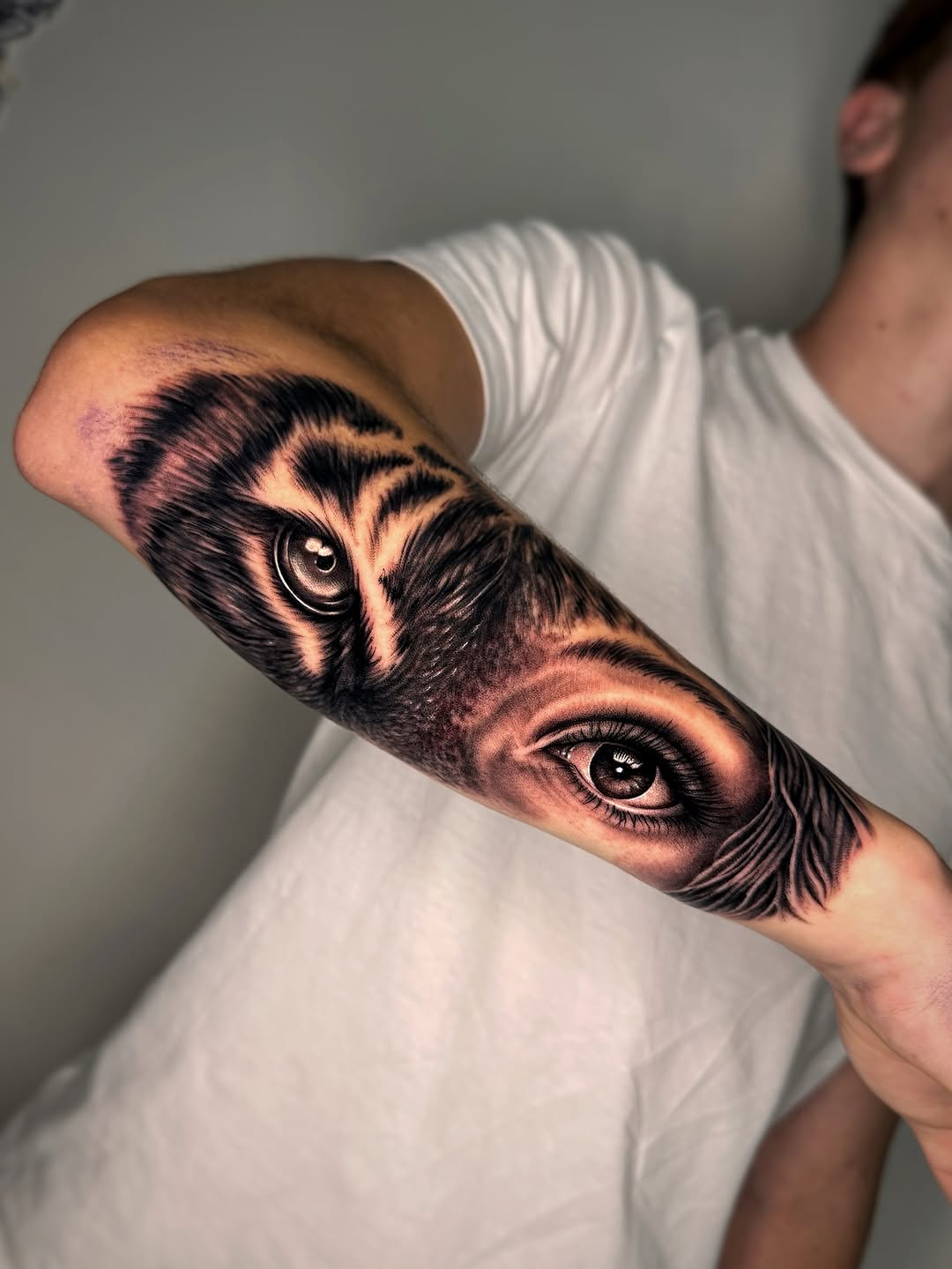 Black and grey realistic owl and human eye tattoo on forearm with detailed feathering and shading.