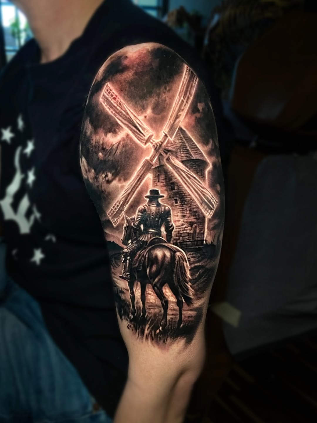 Cowboy on horseback facing a windmill under a dramatic sky, rendered in black and grey shading on upper arm.