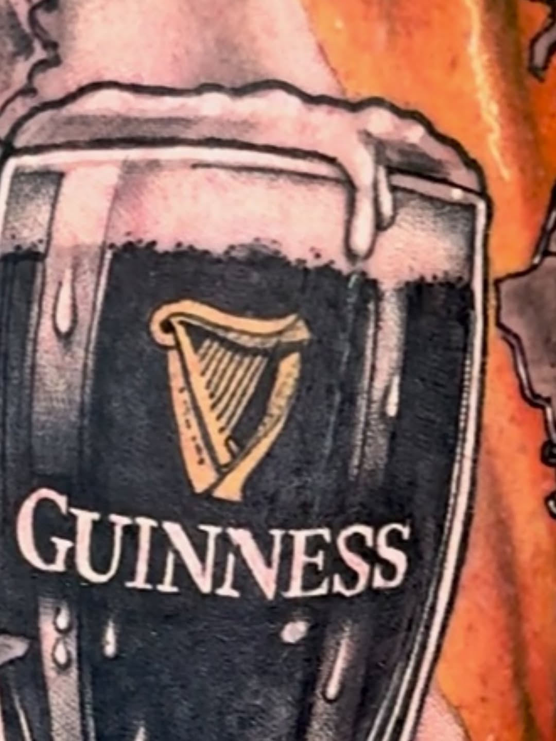 Guinness beer pint glass tattoo with cream head and harp logo in black and grey on arm against orange background.