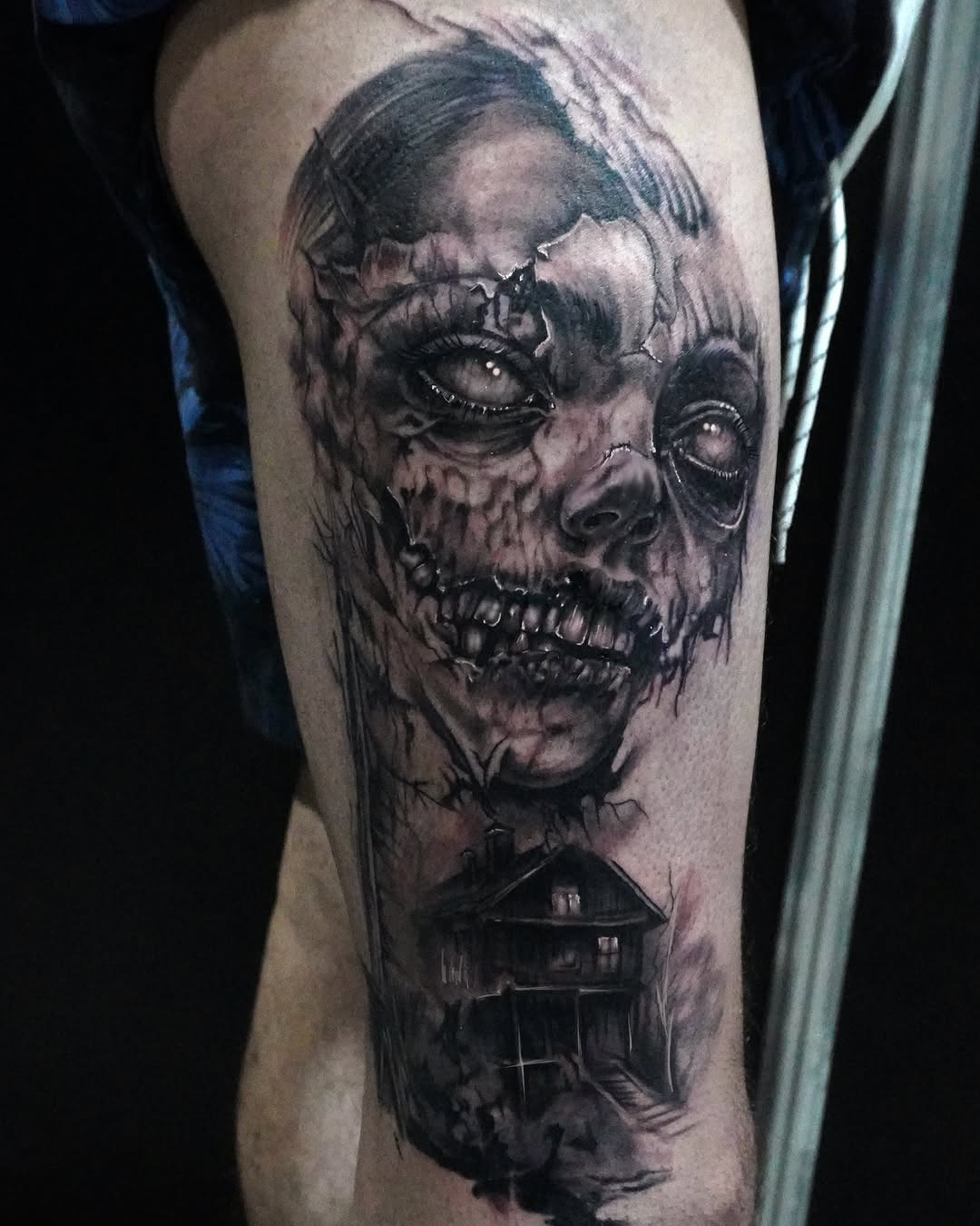 Haunting skull with stitched mouth and glowing eyes above a dark house, rendered in black and grey shading on the thigh.