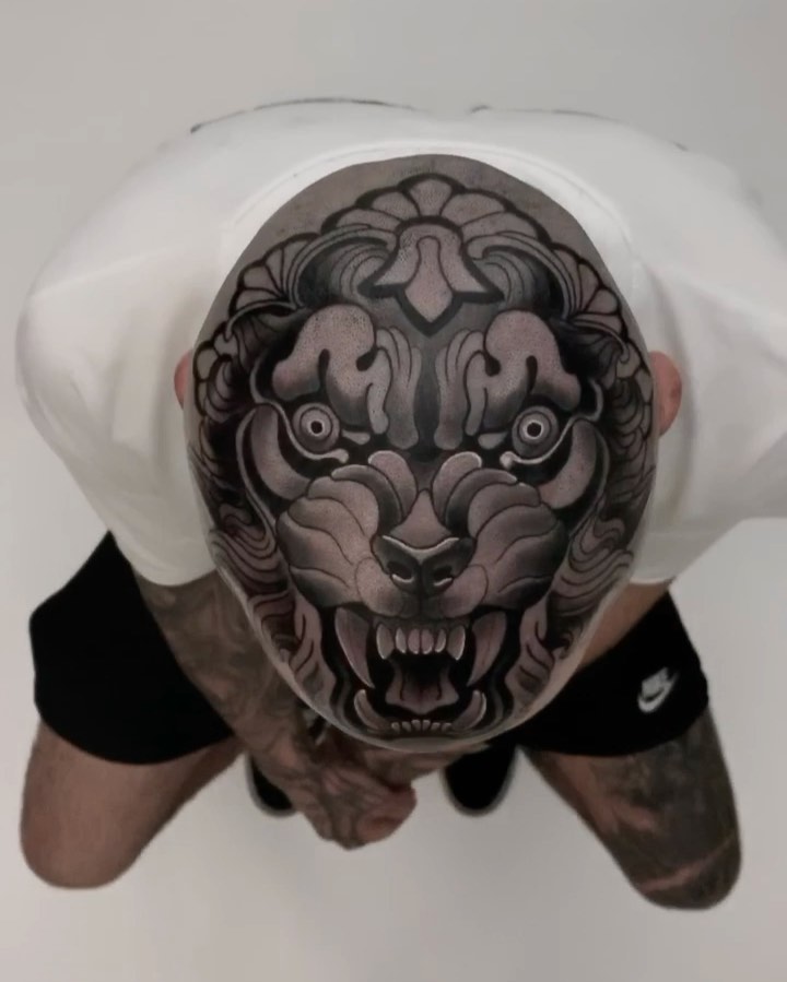Large ornate demon or oni mask tattoo on scalp with intricate shading, grey and black tones, person viewed from above wearing white head wrap.