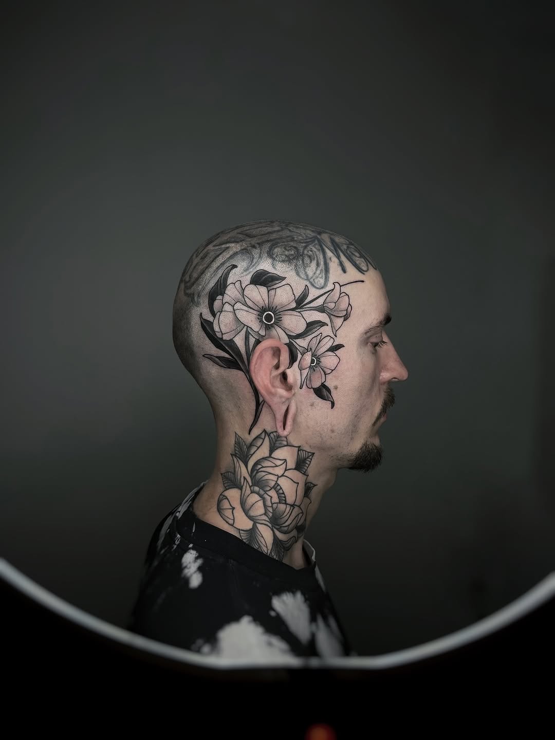 Profile view of man with black and grey floral tattoos covering head, neck, and upper chest featuring detailed flowers with soft shading.