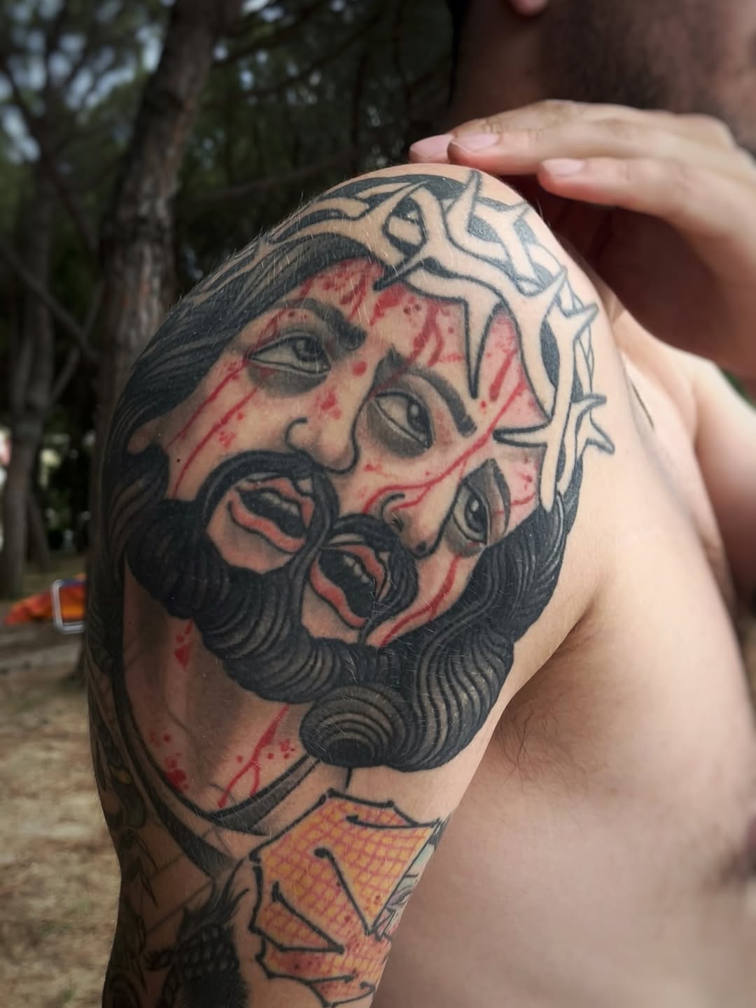 Bearded male face with ornate headpiece and red accents on upper arm, rendered in black and grey with soft shading.