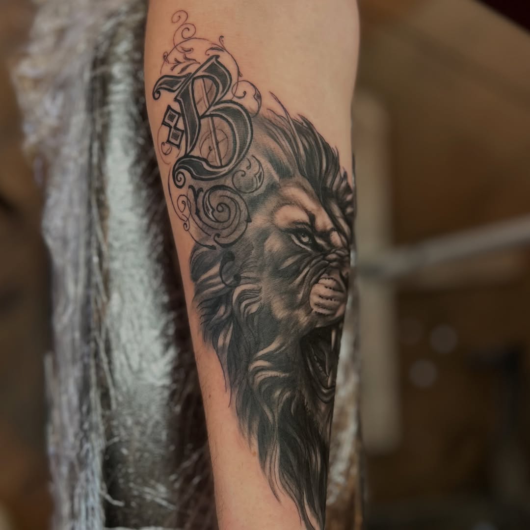 Black and grey portrait tattoo of a bearded man's face with ornate decorative scrollwork above on the forearm.
