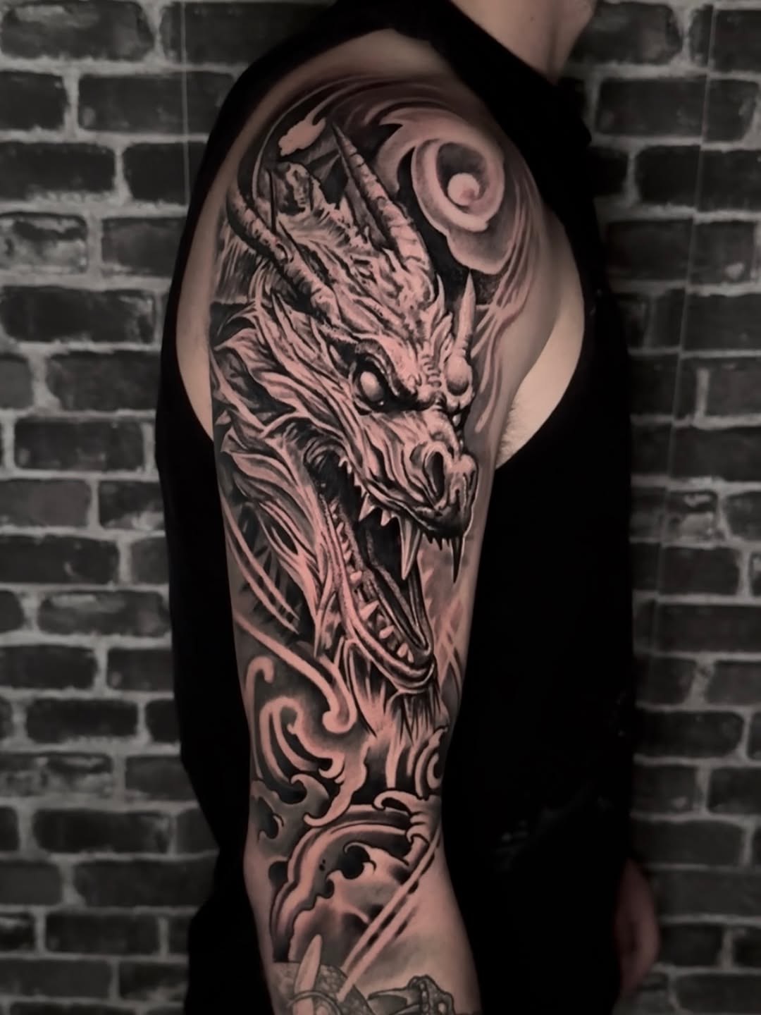 Black and grey dragon head tattoo with ornate scrollwork on upper arm against brick background.