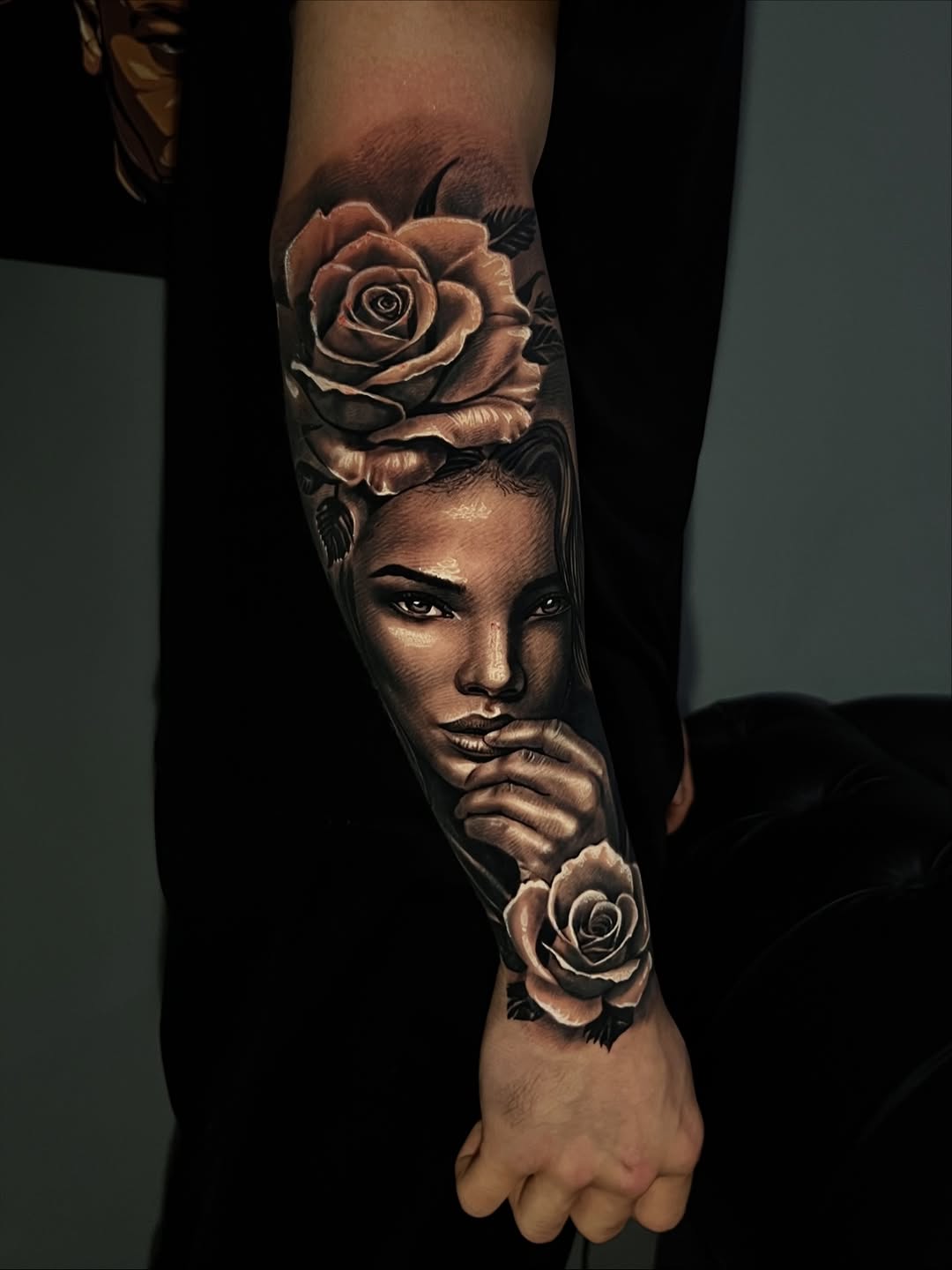 Portrait of a woman's face with roses framing it, rendered in monochrome black and grey on forearm.