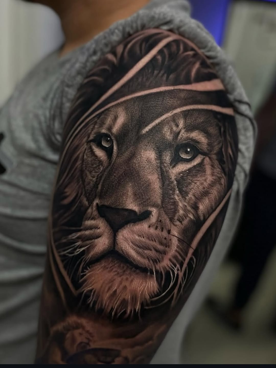 Photorealistic lion head portrait with geometric line frame on upper arm, monochrome black and grey shading.