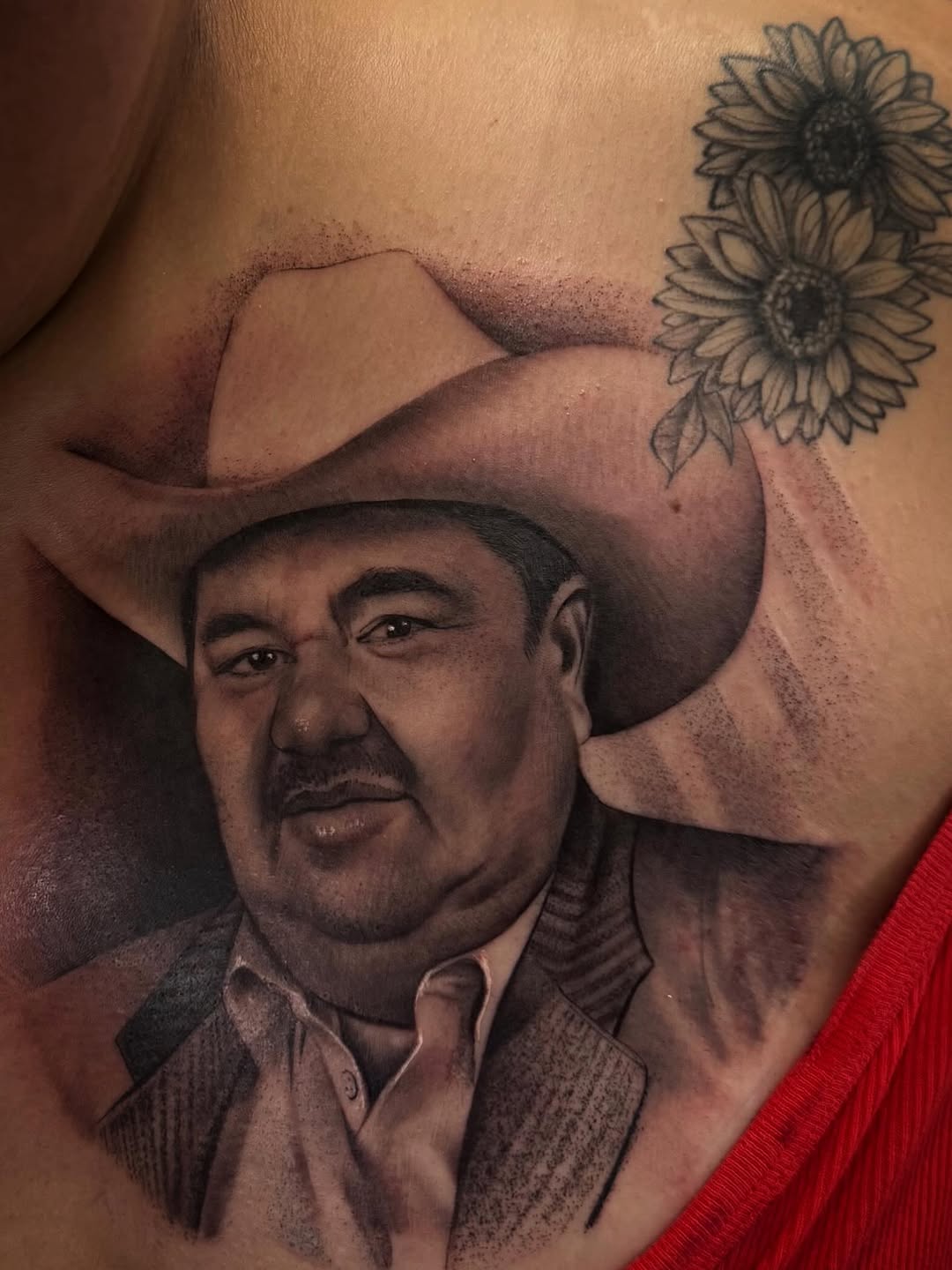 Black and grey portrait tattoo of a man in a cowboy hat and jacket, with sunflowers above, on torso skin.