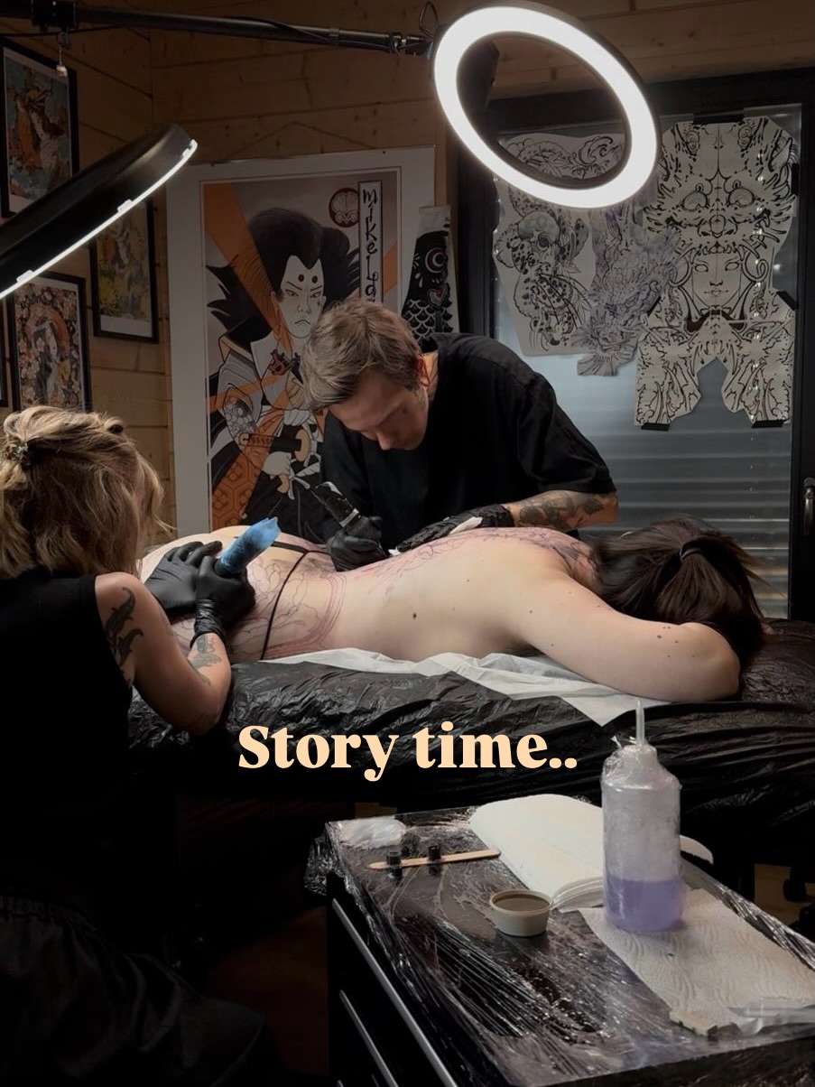 Tattoo artist applying black and grey ink to client's back during session in studio with ring light overhead.