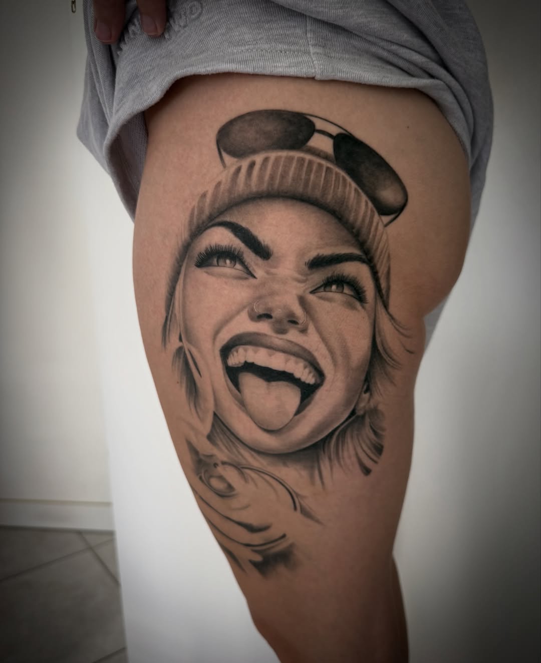 Smiling woman's face with beanie hat tattooed on thigh in black and grey with soft shading and realistic details.