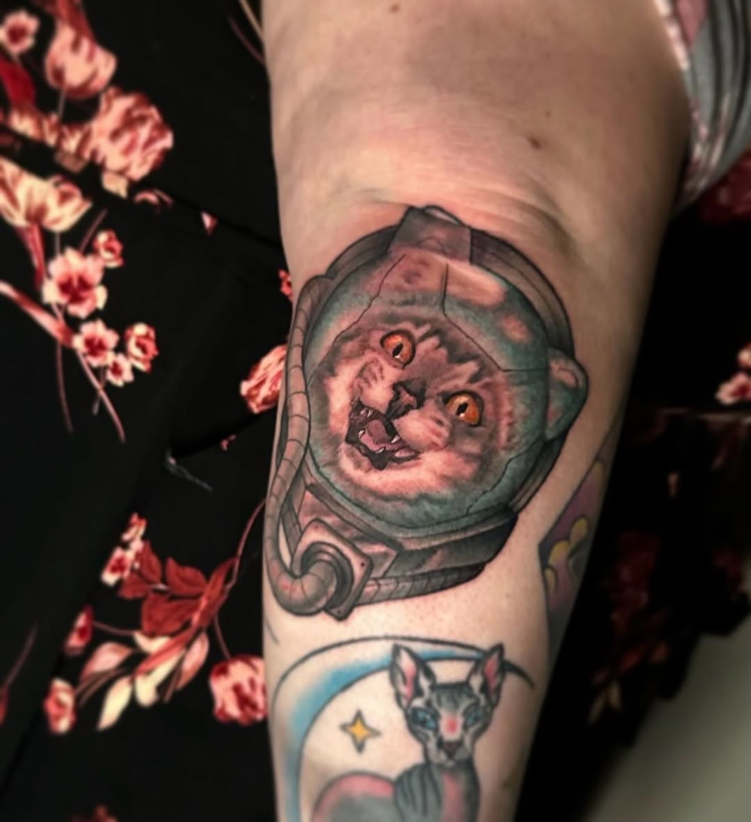 Cat's face inside an astronaut helmet with smooth grey shading on upper arm, with a small celestial cat design below in blue and grey tones.