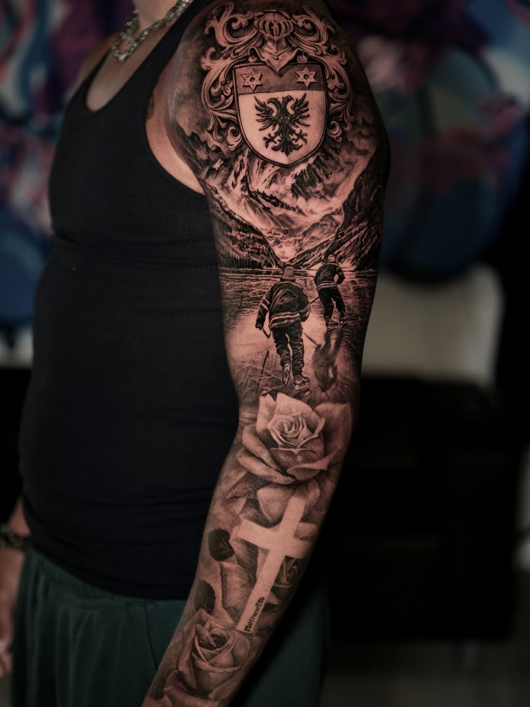 Full sleeve tattoo featuring a heraldic coat of arms, mountainous landscape, astronauts, and roses in black and grey shading on upper arm.