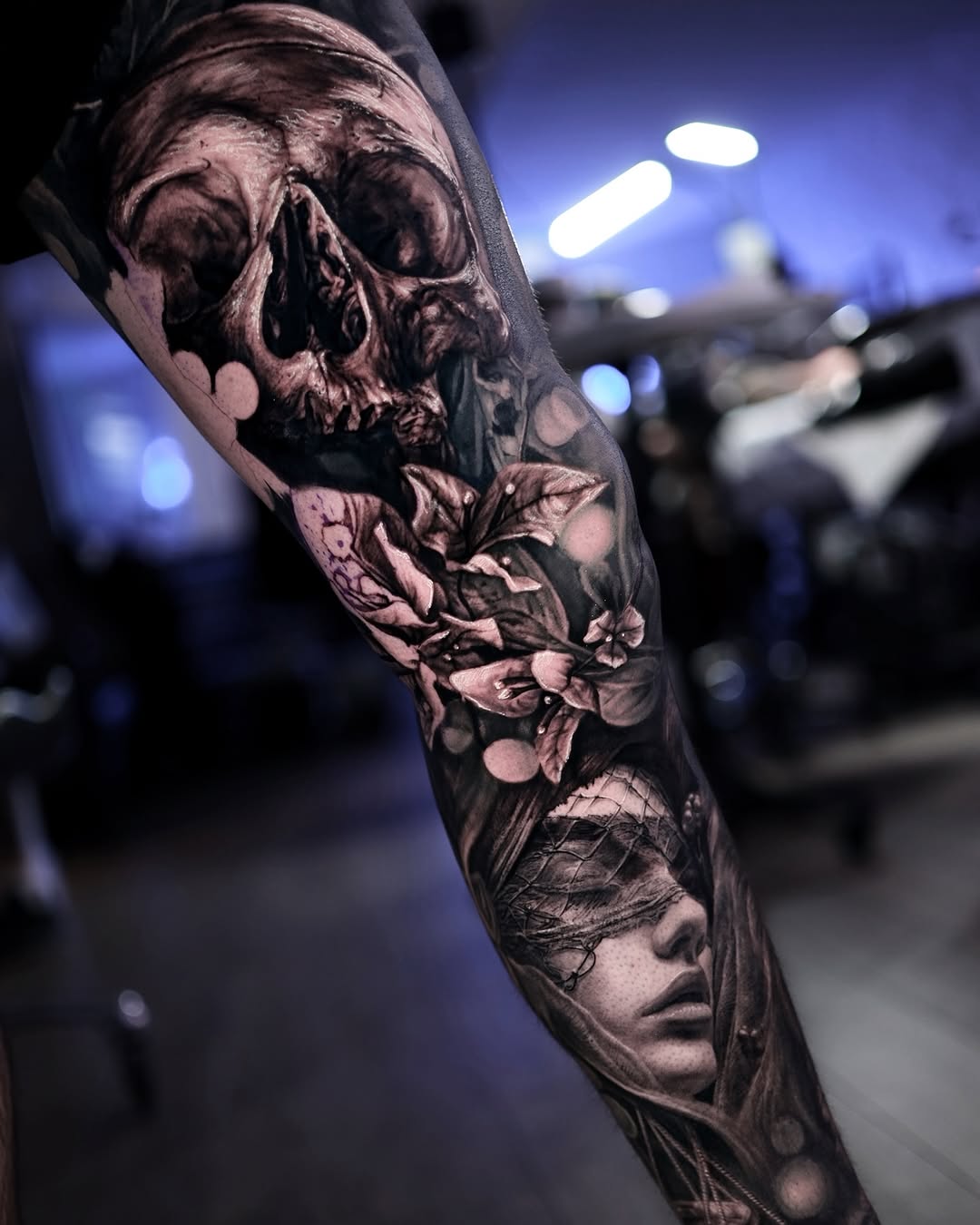 Sleeve tattoo featuring a skull at top, flowering botanicals in middle, and a face portrait at bottom in black and grey shading on arm.