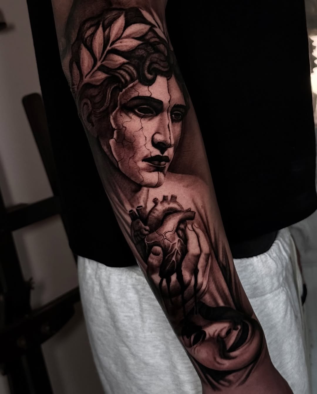 Black and grey portrait tattoo of a classical male face with laurel wreath and anatomical heart on forearm.