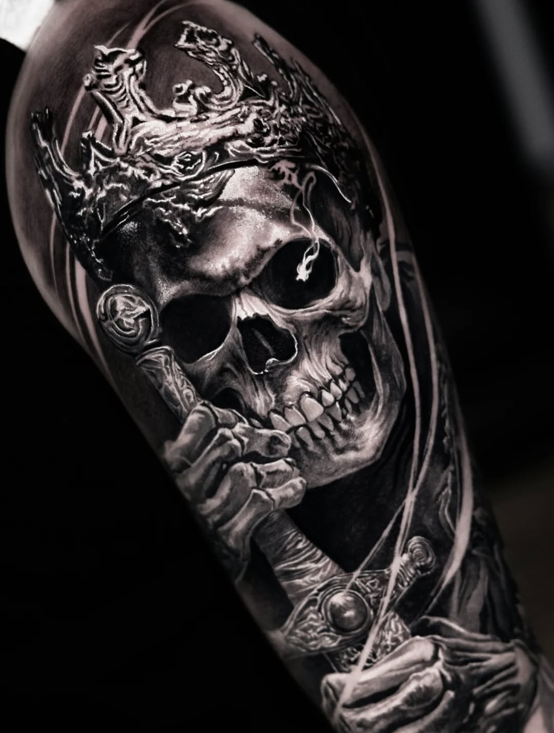 Ornate skull with decorative flourishes and scrollwork, rendered in monochromatic black and grey with photorealistic shading on upper arm.