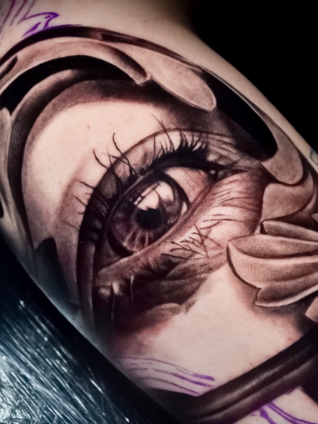 Detailed black and grey eye tattoo on shoulder with dramatic shading, realistic lashes, and flowing fabric elements surrounding it.
