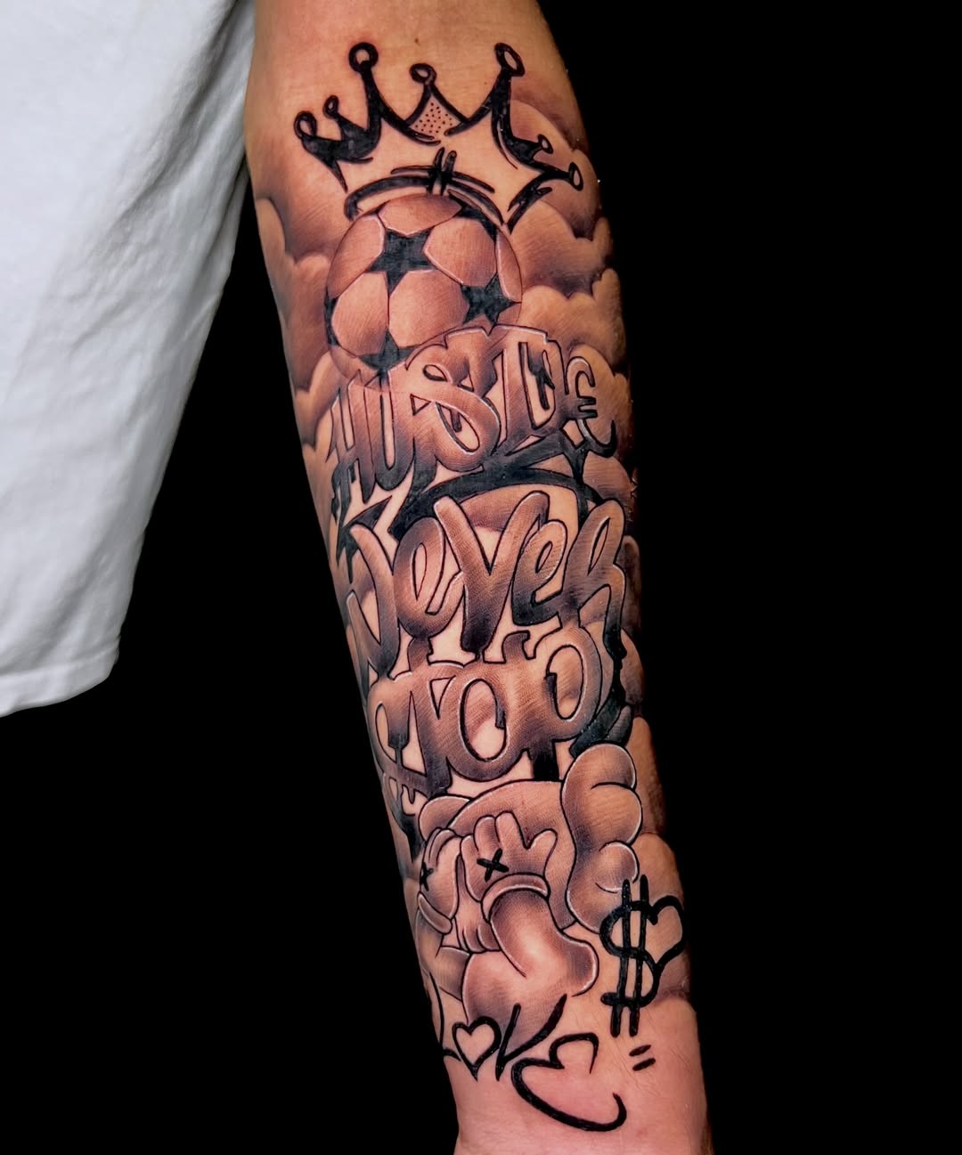 Forearm tattoo featuring bubble letters spelling text with crown, stars, dollar sign, and heart symbols in black and grey shading.