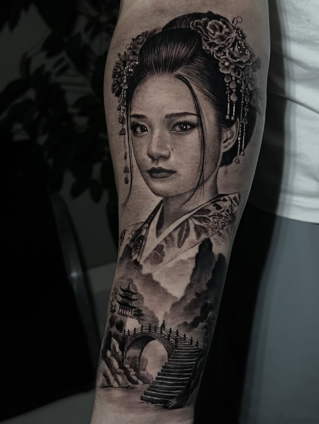 Black and grey portrait tattoo of an Asian woman in traditional dress with ornate hair accessories and dangling earrings, integrated with a landscape featuring pagodas and bridge below on the leg.