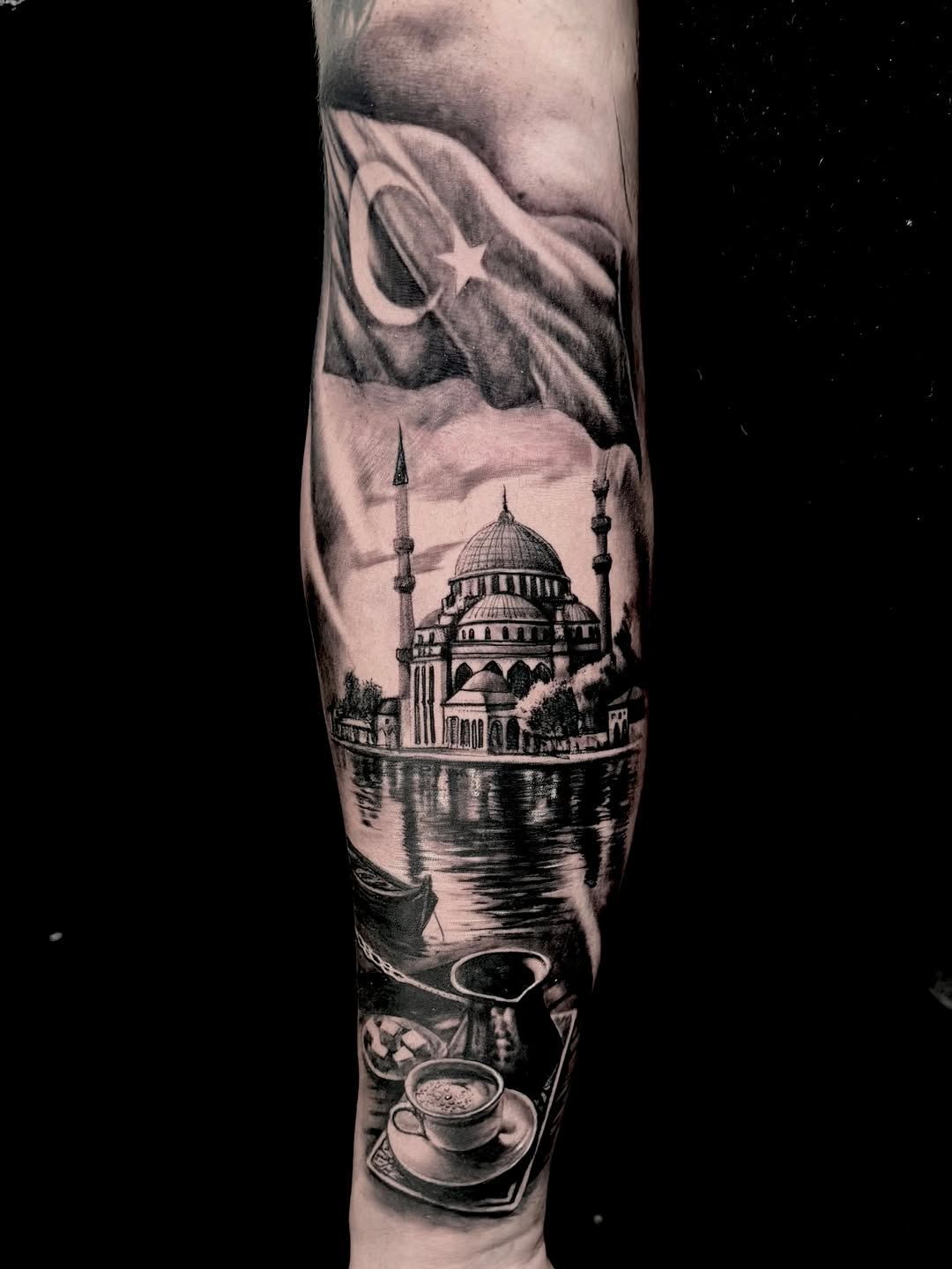 Black and grey forearm sleeve tattoo featuring a mosque with minarets reflected in water, Pakistani flag, and teacups in foreground.
