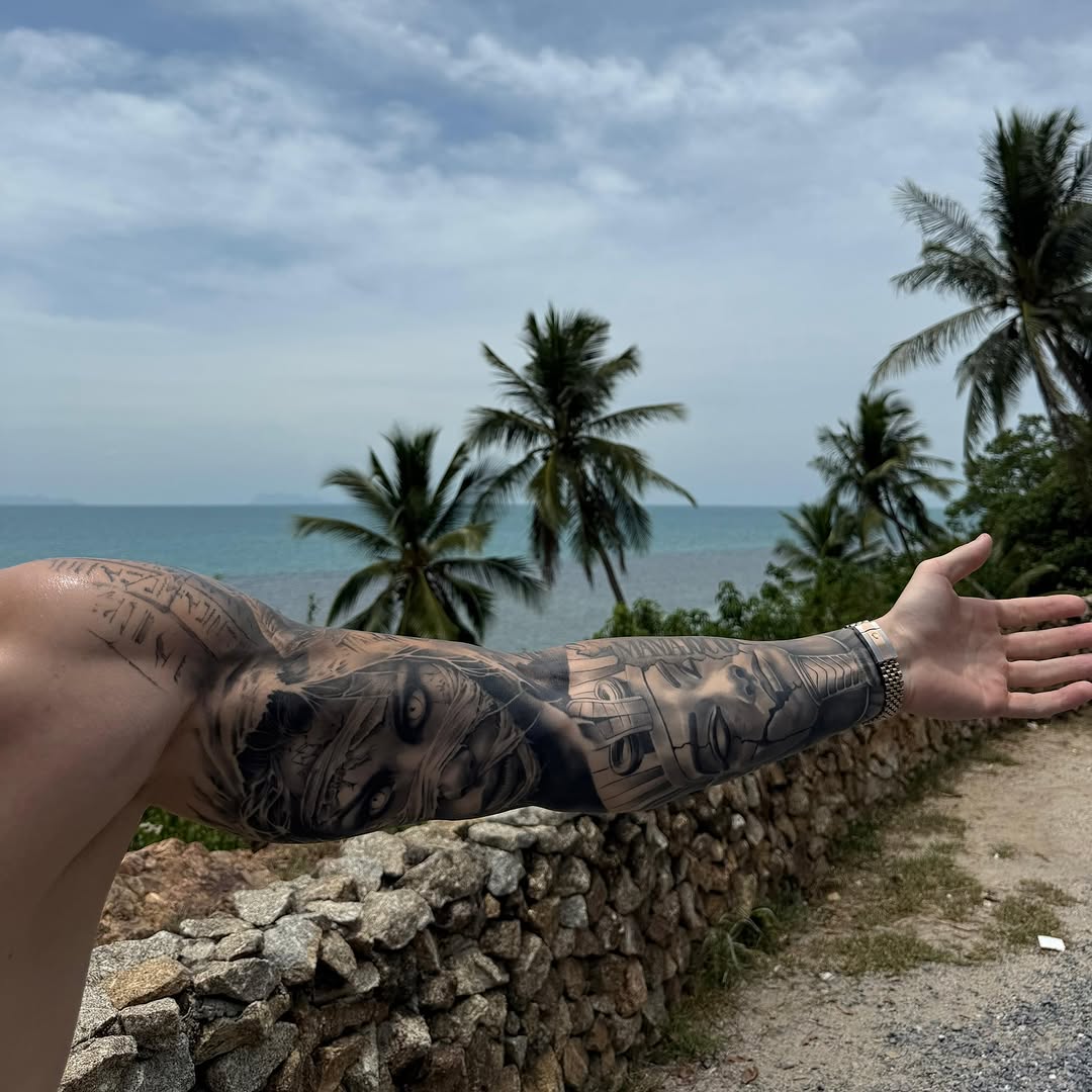 Tattooed arm extended toward ocean with palm trees, featuring black and grey detailed portrait and geometric designs on forearm.