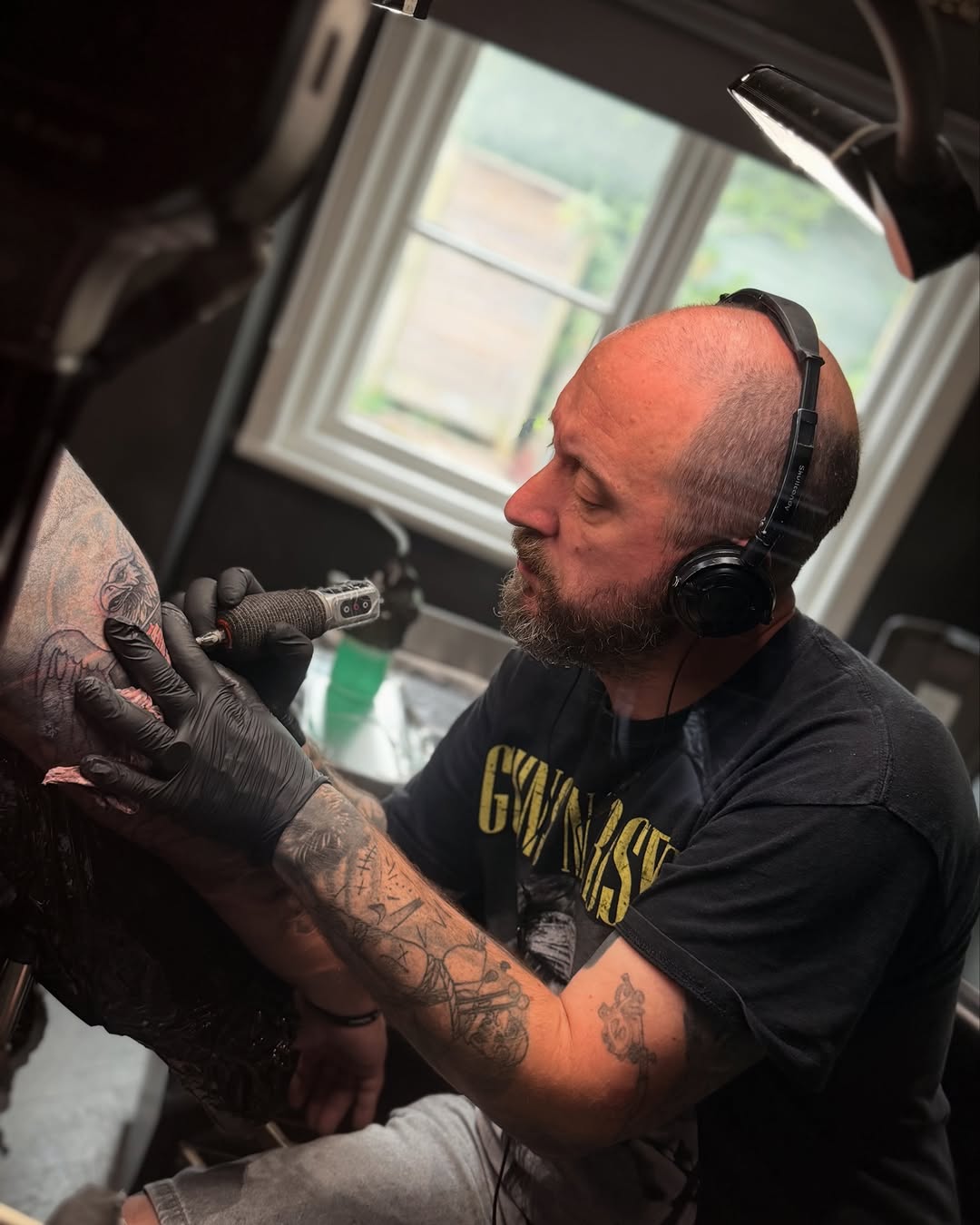 Tattoo artist wearing headphones applies black and grey ink to client's arm in bright studio with window backdrop.