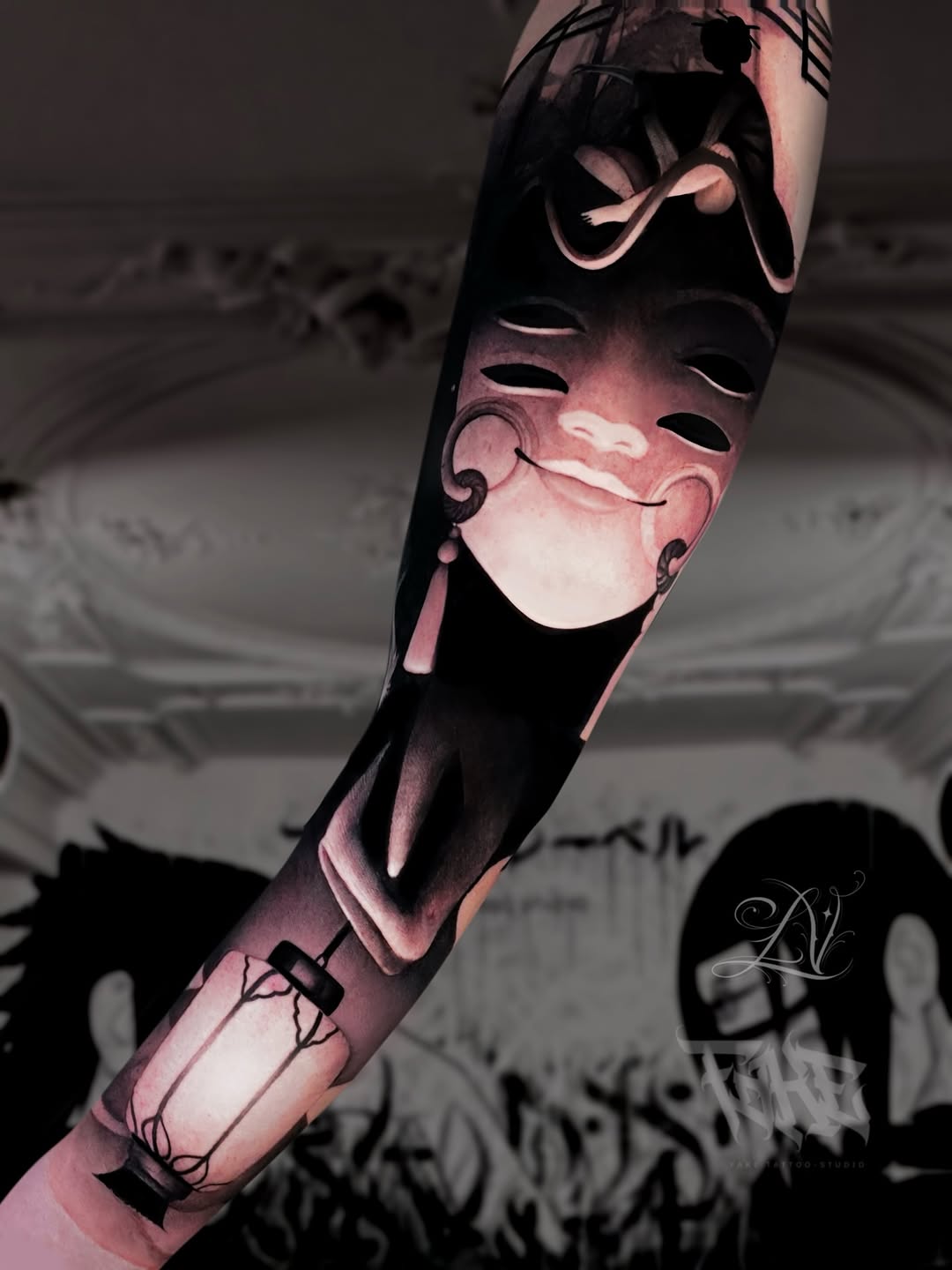 Forearm sleeve tattoo of a smiling theatrical mask with tassels above a lantern and figures, rendered in black and grey shading.