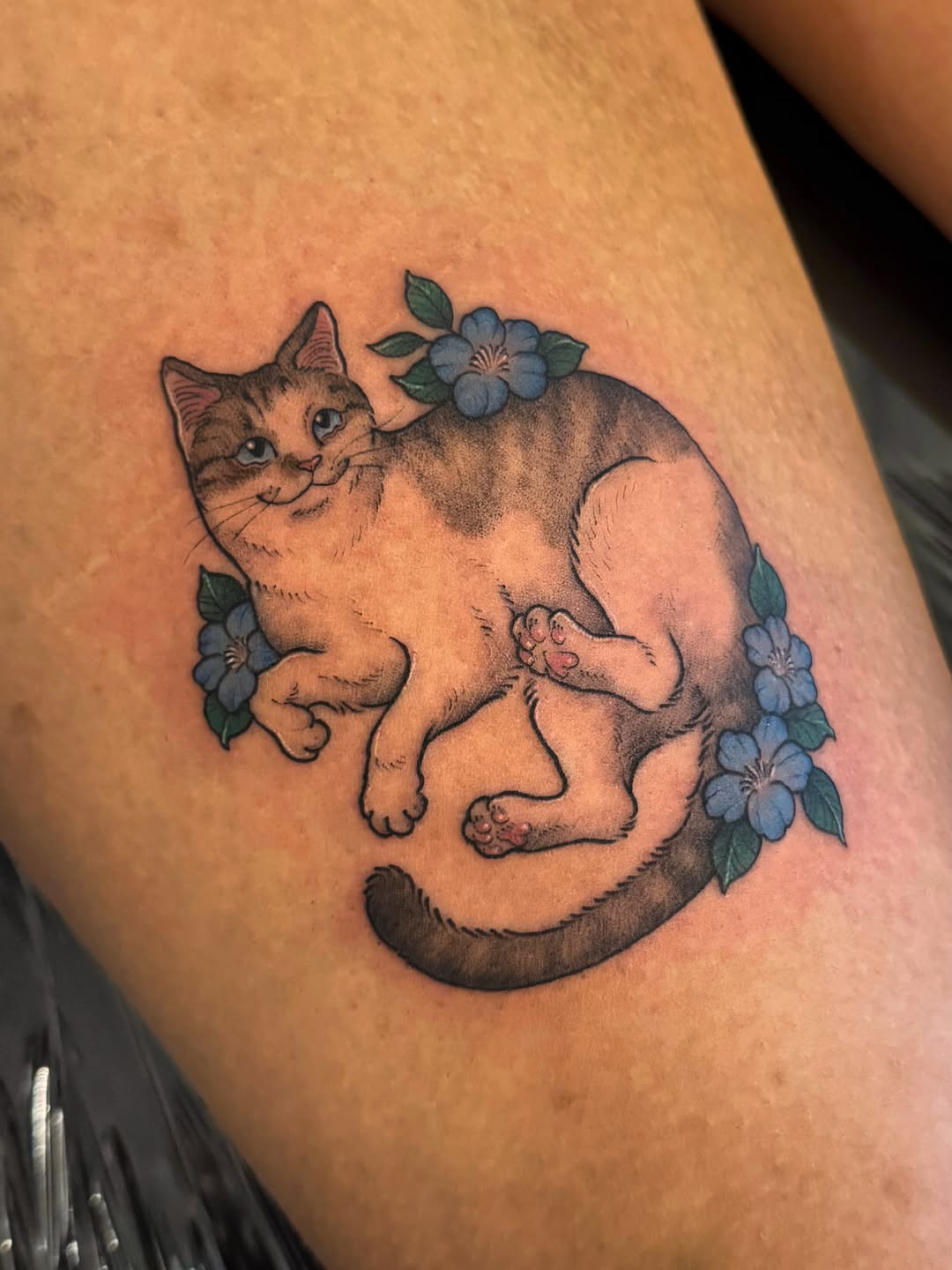 Tabby cat with blue flowers in its fur and tail, sitting position, on upper arm with soft black and grey shading.