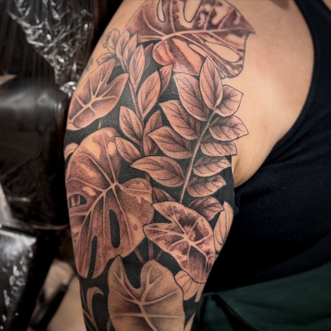 Black and grey botanical tattoo featuring layered leaves and flowers with detailed shading on upper arm.