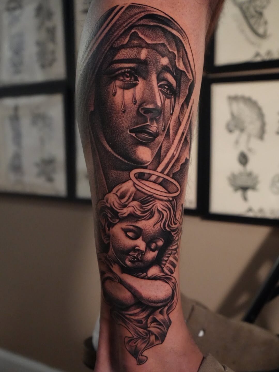Black and grey portrait tattoo on forearm featuring a weeping hooded figure above a cherub with curly hair and halo, detailed shading throughout.