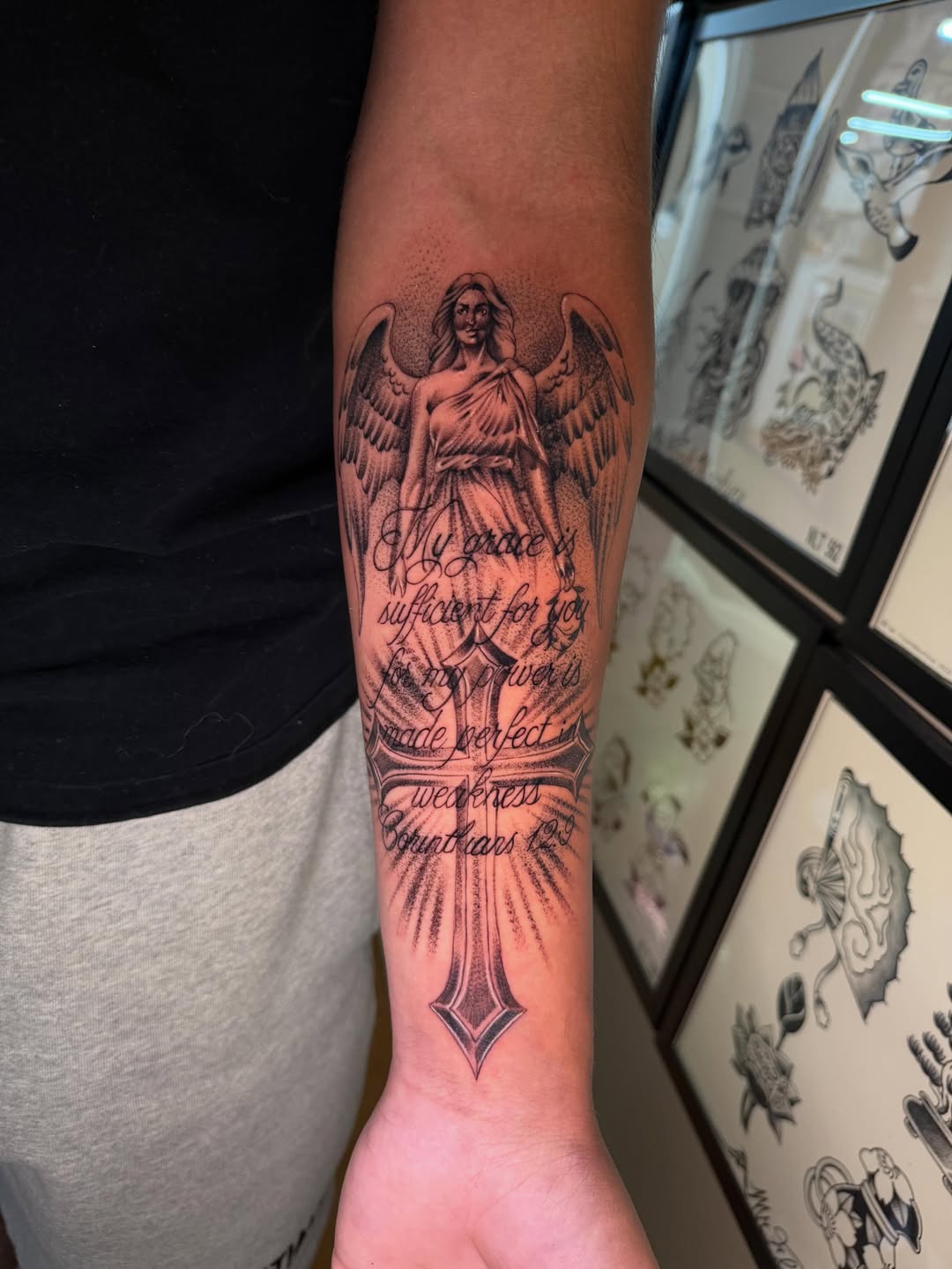 Angel with wings and flowing robes holding a cross, with script text wrapping around the forearm in black and grey.
